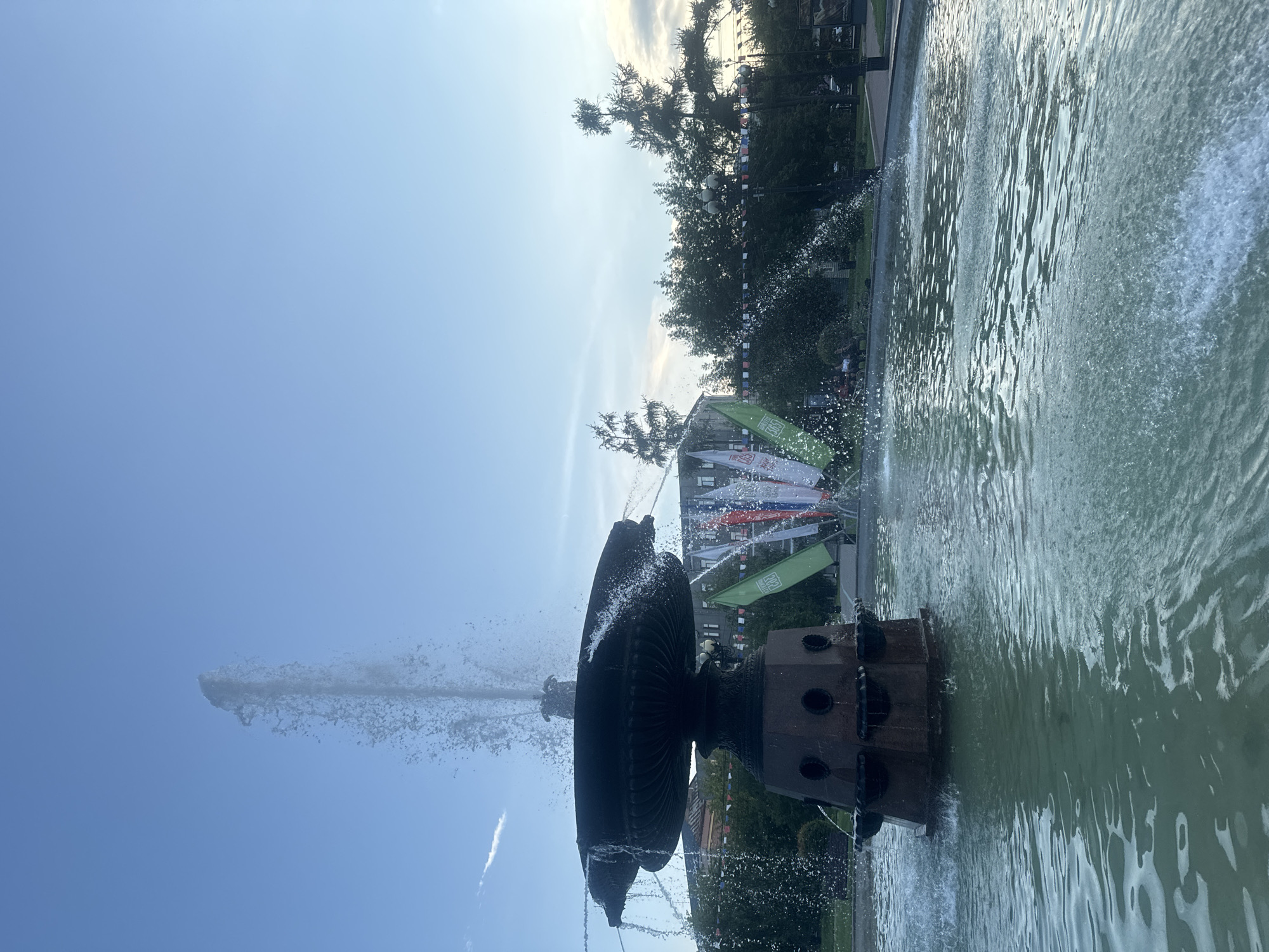 The image shows a fountain with water spraying upwards in a park. In the background, there are trees, buildings, and banners. The sky is blue with some clouds. The fountain is made of dark material and has multiple tiers. The water in the fountain is clear and creates ripples on the surface. There are some people visible in the background, sitting on benches or walking. The overall scene suggests a public outdoor space. This fountain is located in the Kirov Square in Irkutsk, Russia.