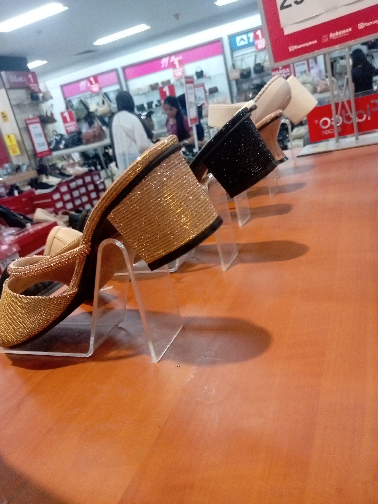 The image captures a display of women's sandals inside a department store in Batam, Indonesia. In the foreground, a gold-colored sandal with intricate beading is prominently displayed on a clear acrylic stand. Behind it, a black sandal with a similar textured design is also on display, followed by a neutral-colored sandal. The sandals are arranged on a wooden surface, likely a display table.
In the background, the store environment is visible. Shelves are stocked with various bags and other merchandise. Several shoppers and store employees are present, though their faces are not clearly discernible. Signs with pricing and promotional information are visible, including one that says "Robinson Ramayana," a known department store chain in Indonesia. The overall lighting suggests an indoor commercial setting.