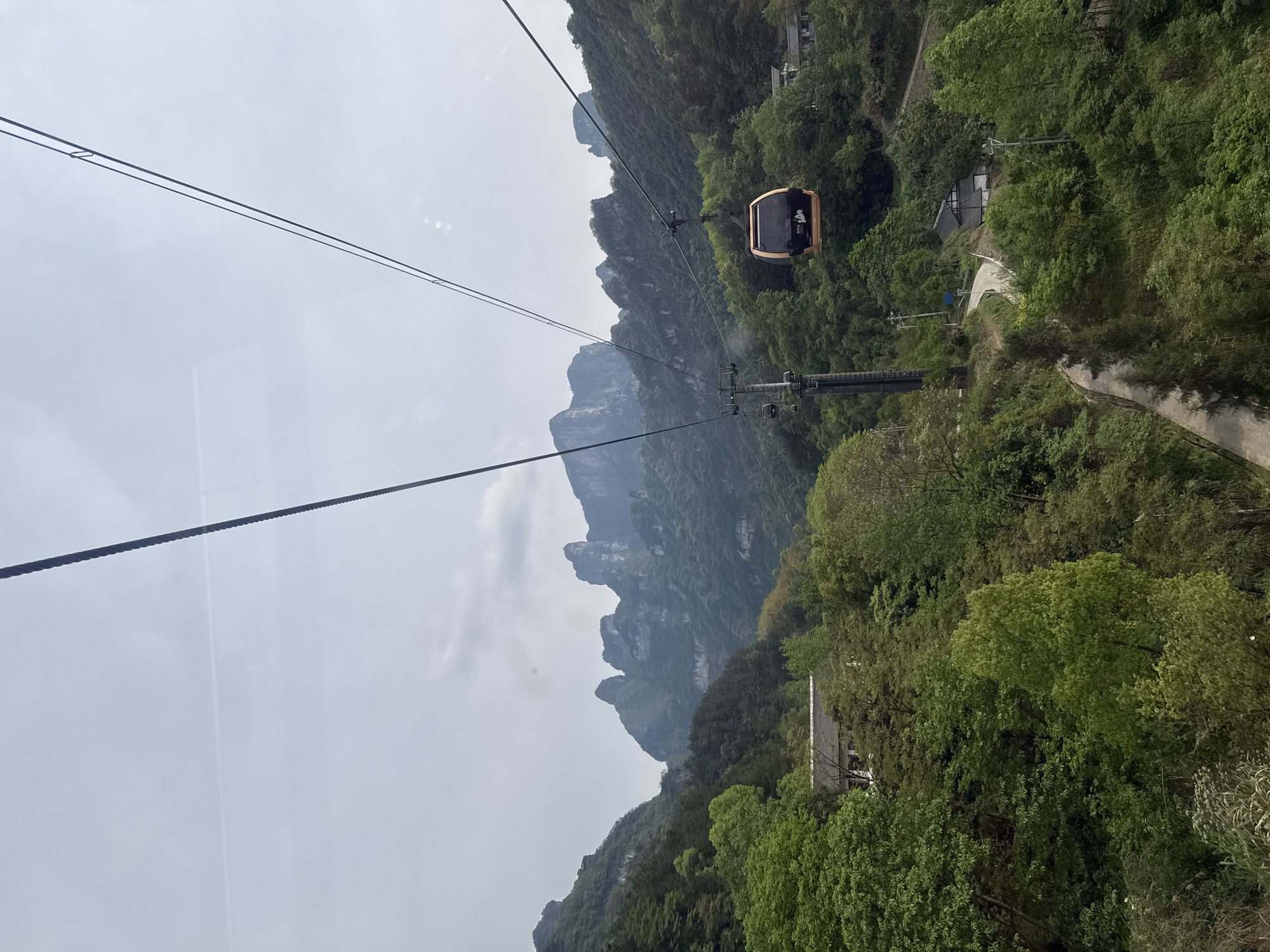 The image shows a cable car suspended high above a lush, mountainous landscape in Yongding District, China. The foreground features the cable lines leading to a tan-colored gondola moving along the cable. A support tower for the cable car is visible below the gondola, surrounded by dense green foliage. In the background, jagged mountain peaks rise into a cloudy sky. Part of a road snakes through the trees near a building, suggesting human presence within the natural environment.
