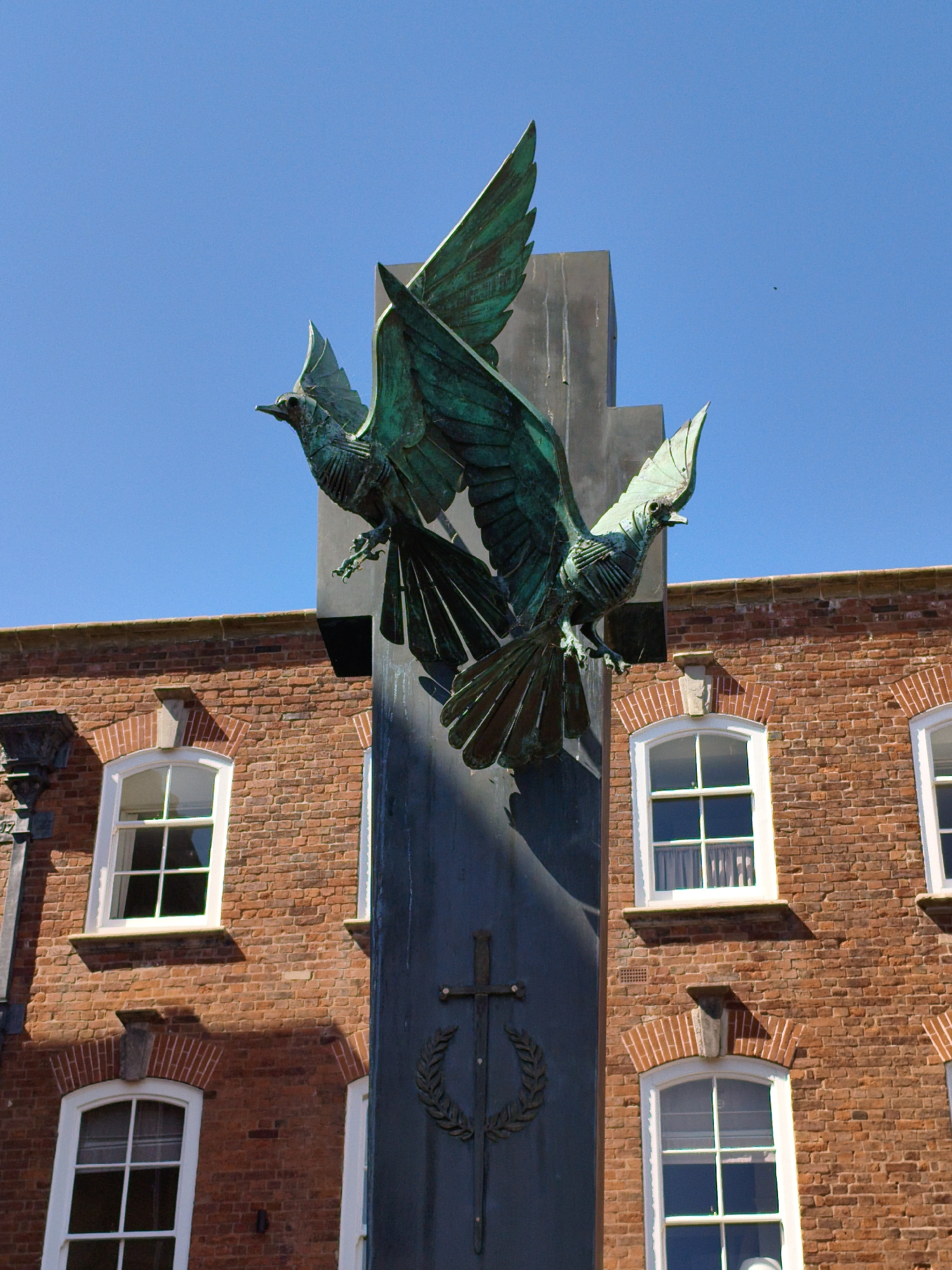 A bronze sculpture of two birds with outstretched wings is mounted on a dark, rectangular column. The birds, with detailed feathers and talons, appear to be in flight, with their heads turned slightly. The column itself features a cross symbol with a laurel wreath. The sculpture is set against a clear blue sky. In the background, a red brick building with white-framed windows is visible. The windows are arranged in rows, and some have white curtains. The building's architecture suggests a historical context. The scene is captured from a low angle, emphasizing the grandeur of the sculpture. This is the Ludlow War Memorial, located in Ludlow, United Kingdom.