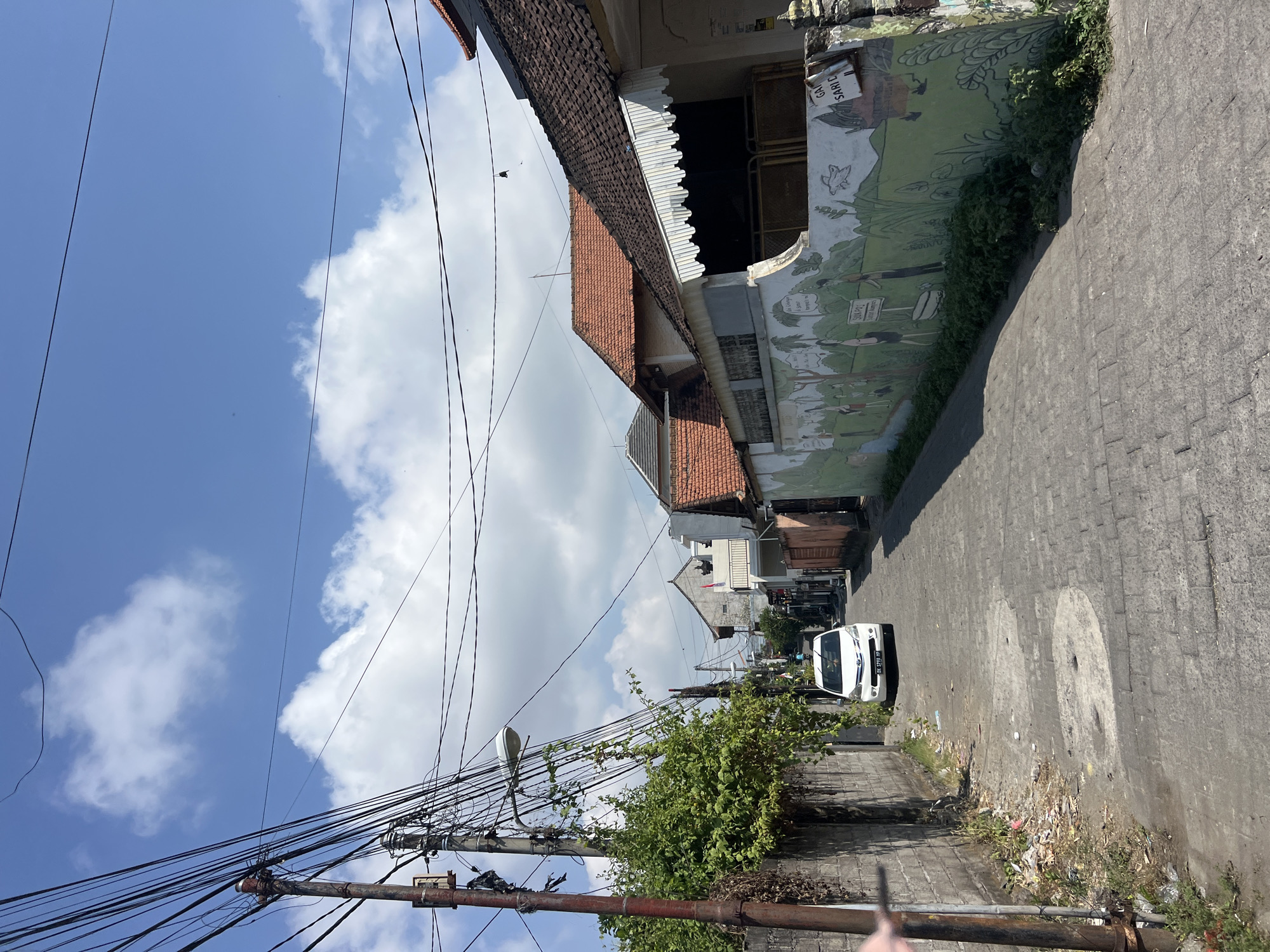 A narrow, paved alleyway runs between buildings in a dense urban environment. A white car is parked in the middle of the alley. The buildings on either side of the alley are residential, with tiled roofs and some of them decorated with murals. The sky is blue with scattered clouds. The scene appears to be in Denpasar, Indonesia, given the architecture and license plate of the car.