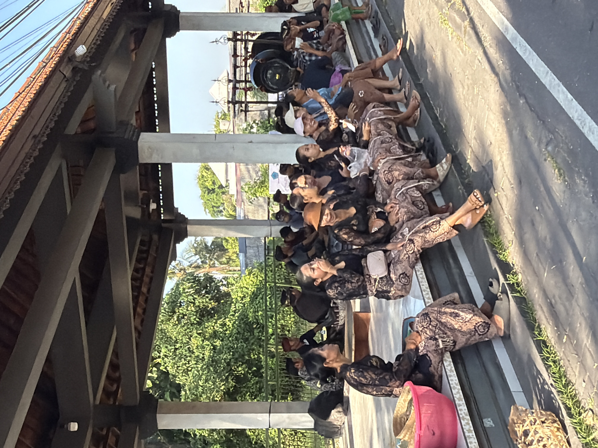 A group of people, mostly women, are seated under a covered pavilion in what appears to be an outdoor gathering in Mengwi, Indonesia. The women are dressed in traditional clothing, featuring patterned skirts and dark tops. Some are engaged in conversation, while others are looking towards the side or holding items. In the foreground, a pink bucket and woven baskets are visible. Behind the seated individuals, trees and buildings can be seen in the background, under a clear sky. The environment suggests a warm, sunny day.