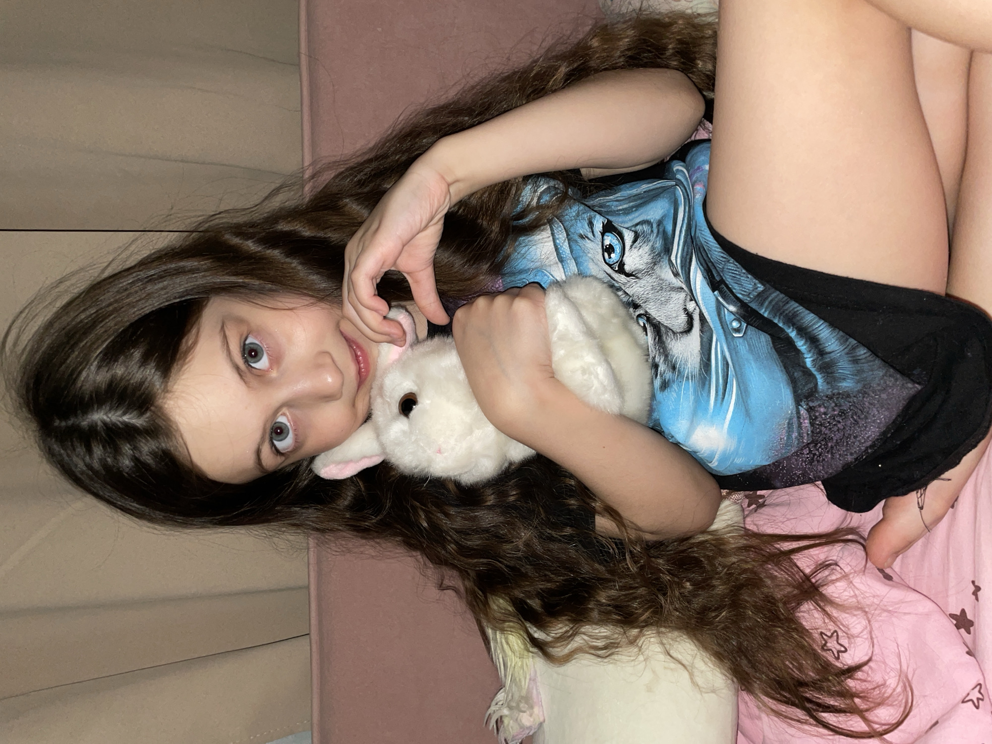 A young girl with long brown hair lies on a pink bed with star patterns, clutching a white stuffed rabbit. She is wearing a blue shirt with a wolf graphic and black shorts. The scene is indoors. The image was not taken in the city of Izhevsk, Russia.