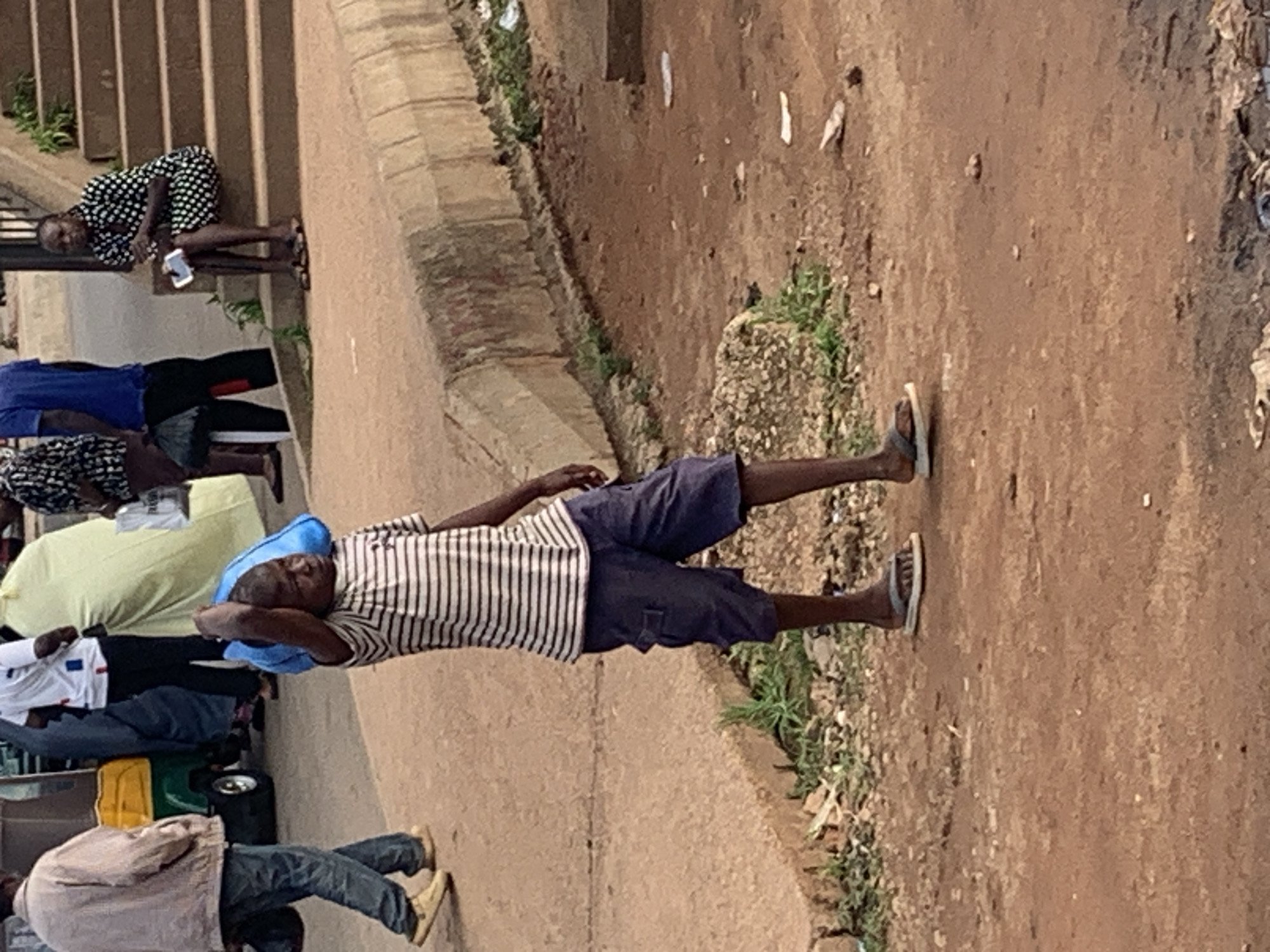 In Gura Top, Nigeria, a young boy wearing a striped t-shirt and dark shorts carries a large blue bag on his head. He walks on a dirt path next to a curb and a grassy area. In the background, a man in a light-colored jacket walks past a green and yellow vehicle. Further back, a woman sits on stairs wearing a black and white polka dot dress, holding a smartphone. Other people are visible in the background, including one in a blue t-shirt and another in a yellow garment. The scene depicts a busy street with various individuals and activities.