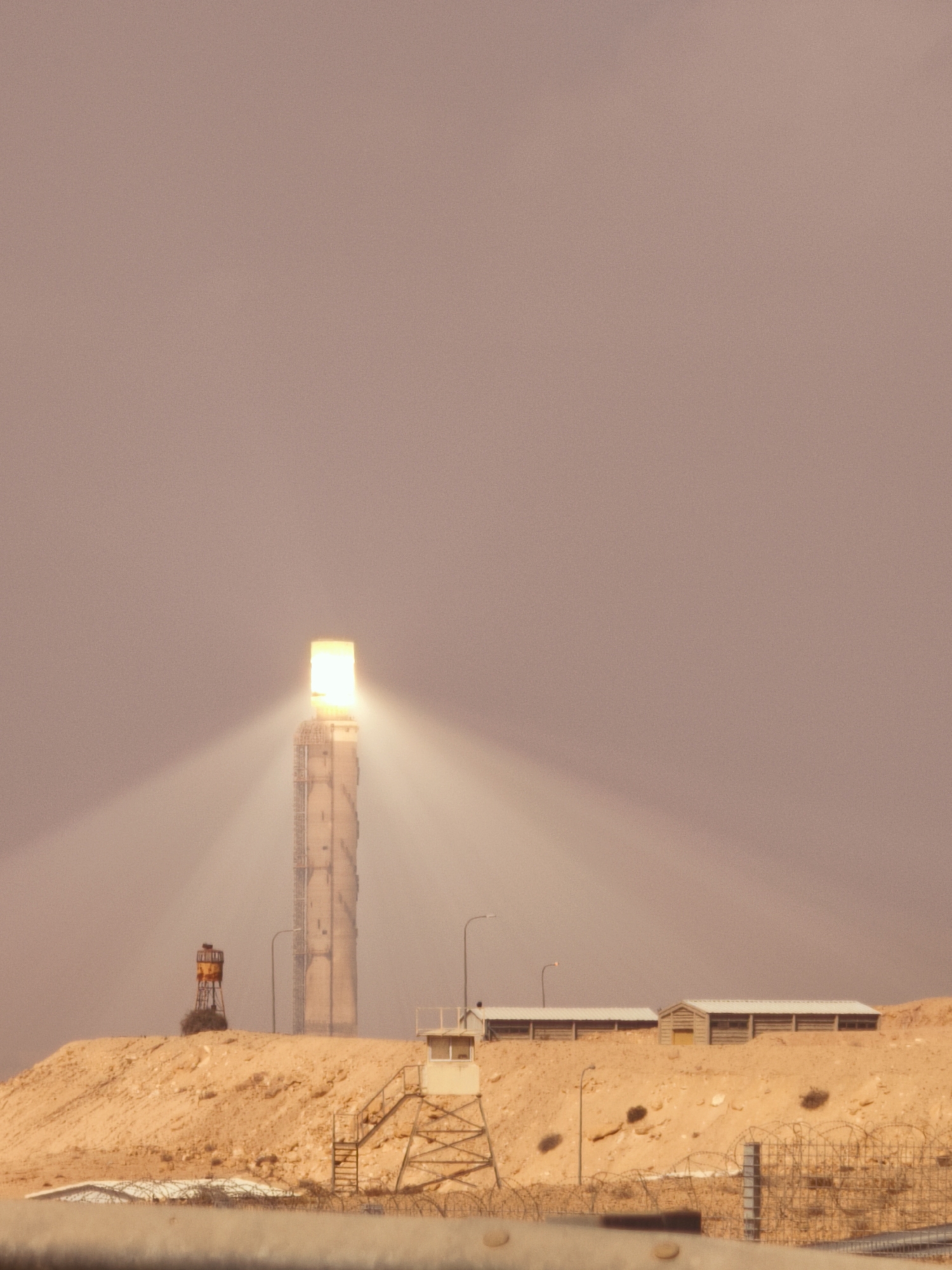 The image depicts a solar thermal power tower in the Negev desert, specifically at the Ashalim Solar Power Station in Israel. The tower, a tall, cylindrical structure, is illuminated at its apex with a bright light source, suggesting it is actively collecting solar energy. Beams of light emanate from the top, creating a hazy glow in the dusty desert atmosphere. In the foreground, the sandy, arid landscape is characterized by low-lying hills. Several smaller structures are visible, including a control booth or guard tower on a hill to the left of the main tower, and two long, low buildings with white roofs further to the right, appearing to be part of the power station's infrastructure. A chain-link fence with barbed wire runs along the very bottom of the frame, indicating a security perimeter. The overall impression is of a large-scale industrial facility in a remote, desert environment.