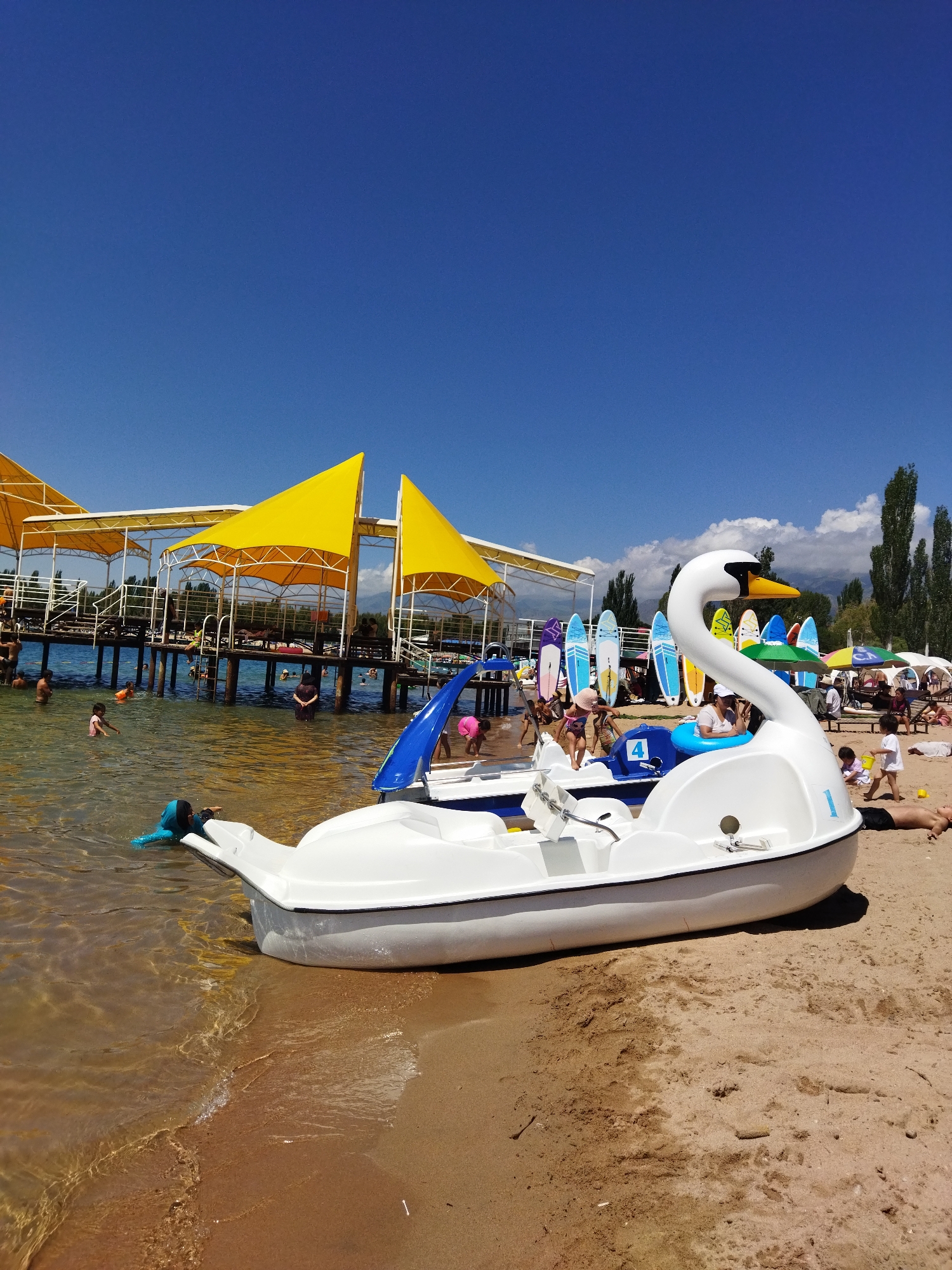 On a sunny day at the beach in Kara-Oy, Kyrgyzstan, a white pedal boat shaped like a swan sits on the sandy shore, its bow gently touching the clear, shallow water. The boat has a yellow beak and black eyes. In the background, a wooden pier extends into the turquoise water, covered by yellow awnings that provide shade. Several people are visible on the pier and in the water, some swimming and others standing. Beyond the pier, a line of tall trees and mountains are visible under a clear blue sky. Along the shoreline, a row of colorful surfboards stands upright, and beach umbrellas are set up. A young child in a blue swimsuit is swimming near the swan boat, while other children play on the sand.