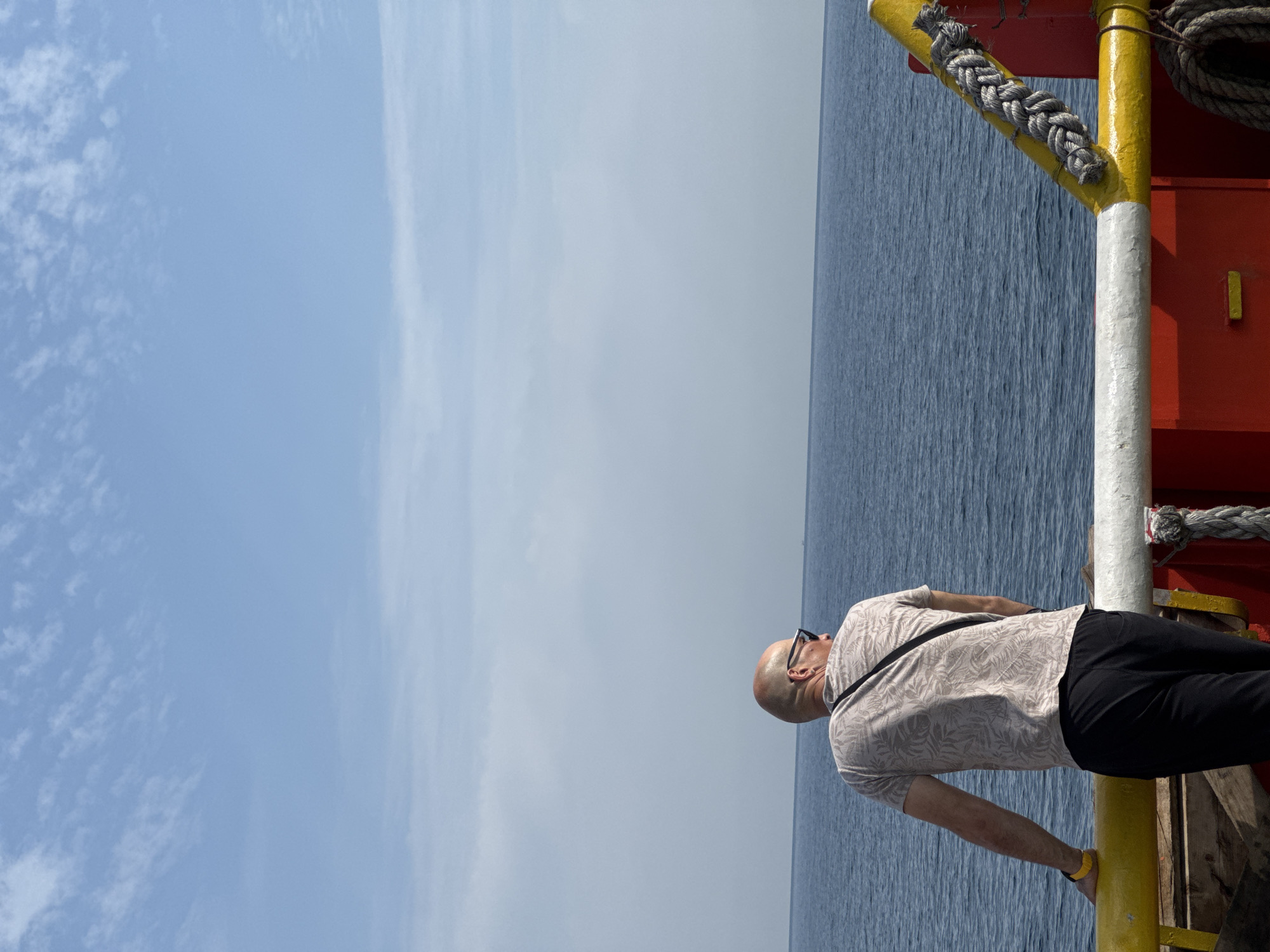 A man, viewed from behind, stands on the deck of a vessel at Marina di Ravenna, Italy, looking out at the sea. He is bald, wears sunglasses and a light-colored, patterned t-shirt, and black pants. He leans his left arm on a yellow railing. The sea is calm and blue, meeting a pale blue sky with wispy clouds. The deck of the vessel is visible in the foreground, with red and yellow painted structures and thick ropes.
