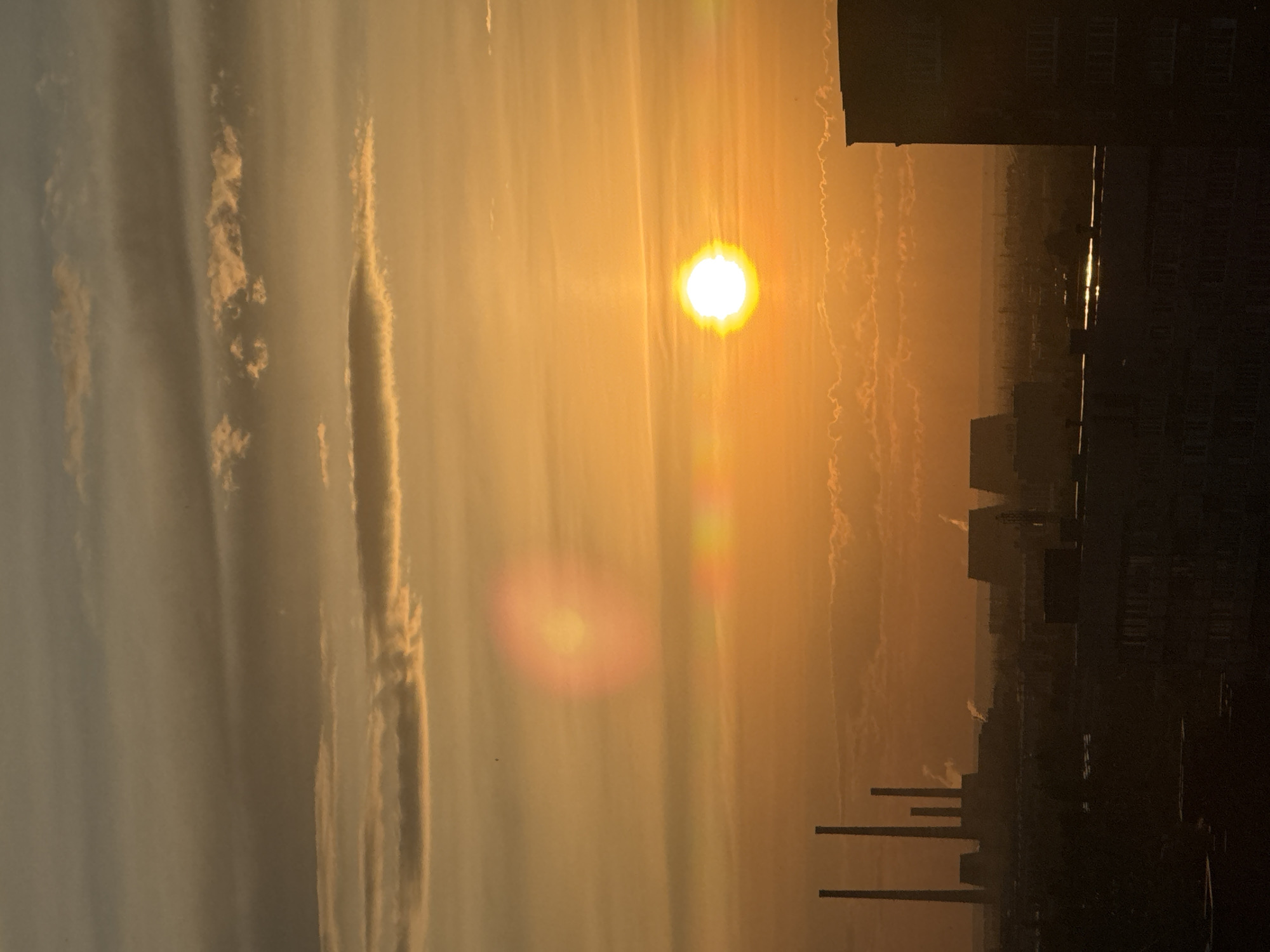 The image depicts a sunset over a city. The sun is a bright, intense orb in the sky, casting a warm orange glow. Several layers of clouds are visible, some illuminated by the sun and others appearing darker. In the distance, a silhouette of an industrial complex with tall smokestacks is present, with faint plumes of smoke rising from them. Towards the foreground and mid-ground, the dark outlines of buildings and structures are visible, suggesting a cityscape. There are lens flares present in the sky, indicating the direct sunlight impacting the camera lens. The overall scene is dominated by the warm colors of the sunset and the silhouettes of industrial and urban elements.