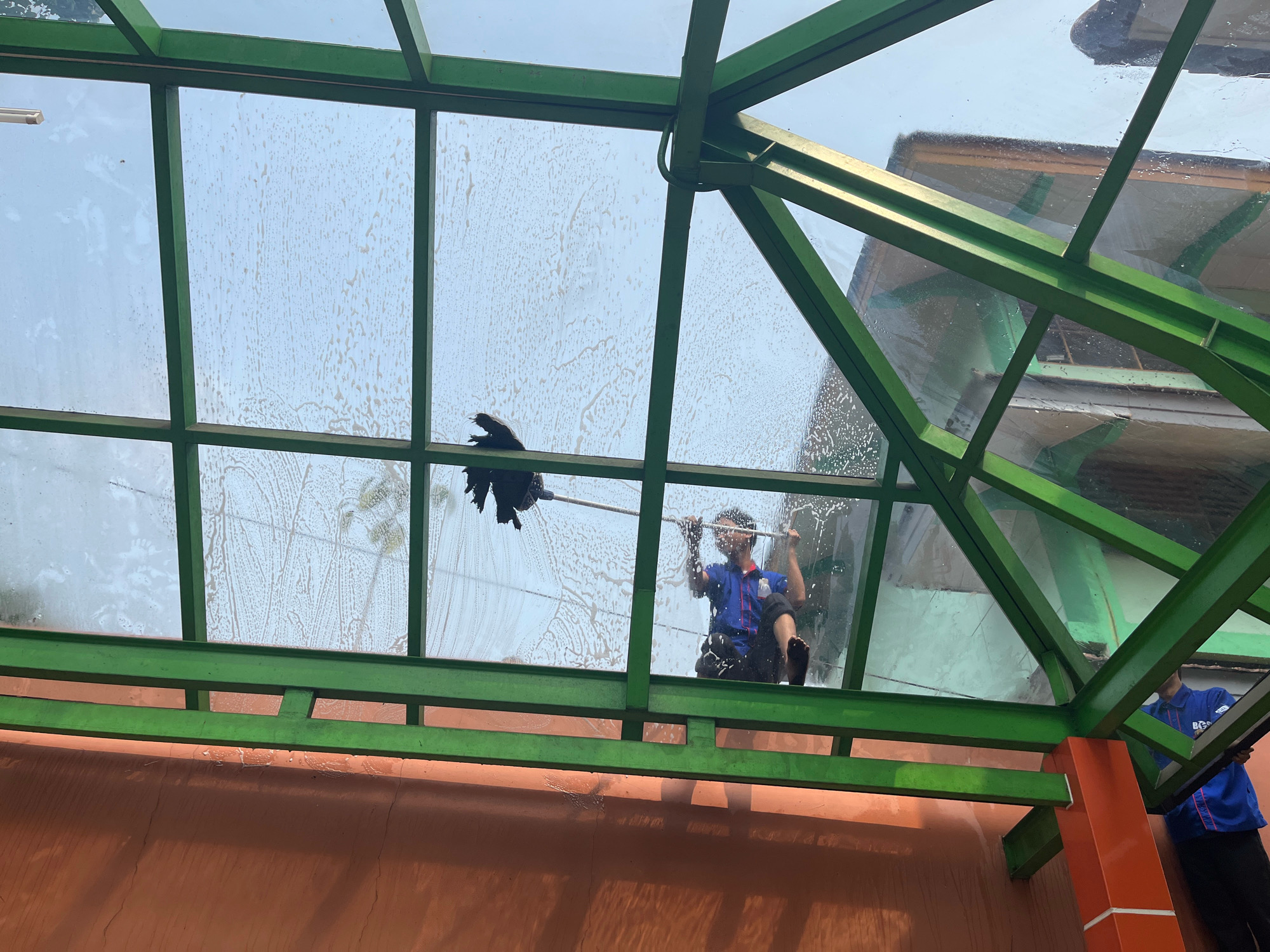 In South Jakarta, Indonesia, a man is cleaning a glass roof. He is wearing a blue uniform and black pants, and he is using a long-handled squeegee. The glass roof is part of a building with a green metal frame and an orange wall. The sky is visible through the glass, and there are some clouds. The glass is dirty and streaky, with soapy water still visible in some areas. Another person in a blue uniform is standing to the right, holding a part of the frame.