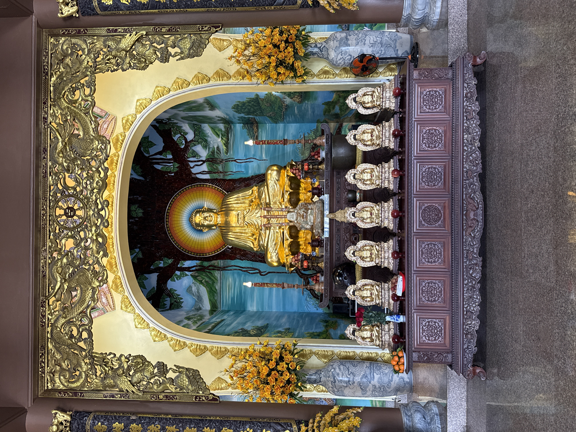 The image depicts the main altar inside Phap Lam Pagoda, located at 574 Ông Ích Khiêm, Hải Châu 2, Hải Châu, Đà Nẵng, Vietnam. The scene is symmetrical and devoid of individuals.
At the center, a large, golden Buddha statue sits in a meditative Dhyana Mudra posture on a tiered pedestal within an arched niche. A multi-colored, rainbow-like mandorla surrounds the Buddha's head. The background of the niche is a painted mural featuring a large tree with visible roots, distant mountains, and a body of water under a light blue sky. The arched niche is framed by an ornate golden filigree structure incorporating prominent dragon and cloud motifs.
Below the main Buddha, a dark, multi-tiered wooden altar table holds ritualistic items. These include two tall, slender, lit candles with reddish-orange flames, small red fruits (appearing as apples or pomegranates), and other small statues. A smaller golden Buddha statue is centrally positioned on this upper altar.
In front of this, a lower, ornately carved dark wood table displays seven smaller, identical golden Buddha statues seated in a row. Offerings on this lower table include a plate of oranges/tangerines on the left, a small blue-and-white patterned vase holding red roses, and additional red fruits.
Flanking the central shrine are two tall, grey-and-white ceramic vases decorated with stylized dragon patterns. Each vase contains a large, dense arrangement of artificial yellow flowers. A small black and orange electric fan is positioned on the floor near the base of the right-hand vase. The shrine area is bordered by dark brown pillars. The floor is polished, dark speckled granite, reflecting the lower parts of the altar. Upper walls and ceiling visible are a solid, muted brown color, with dark carved panels partially visible on the far left and right.