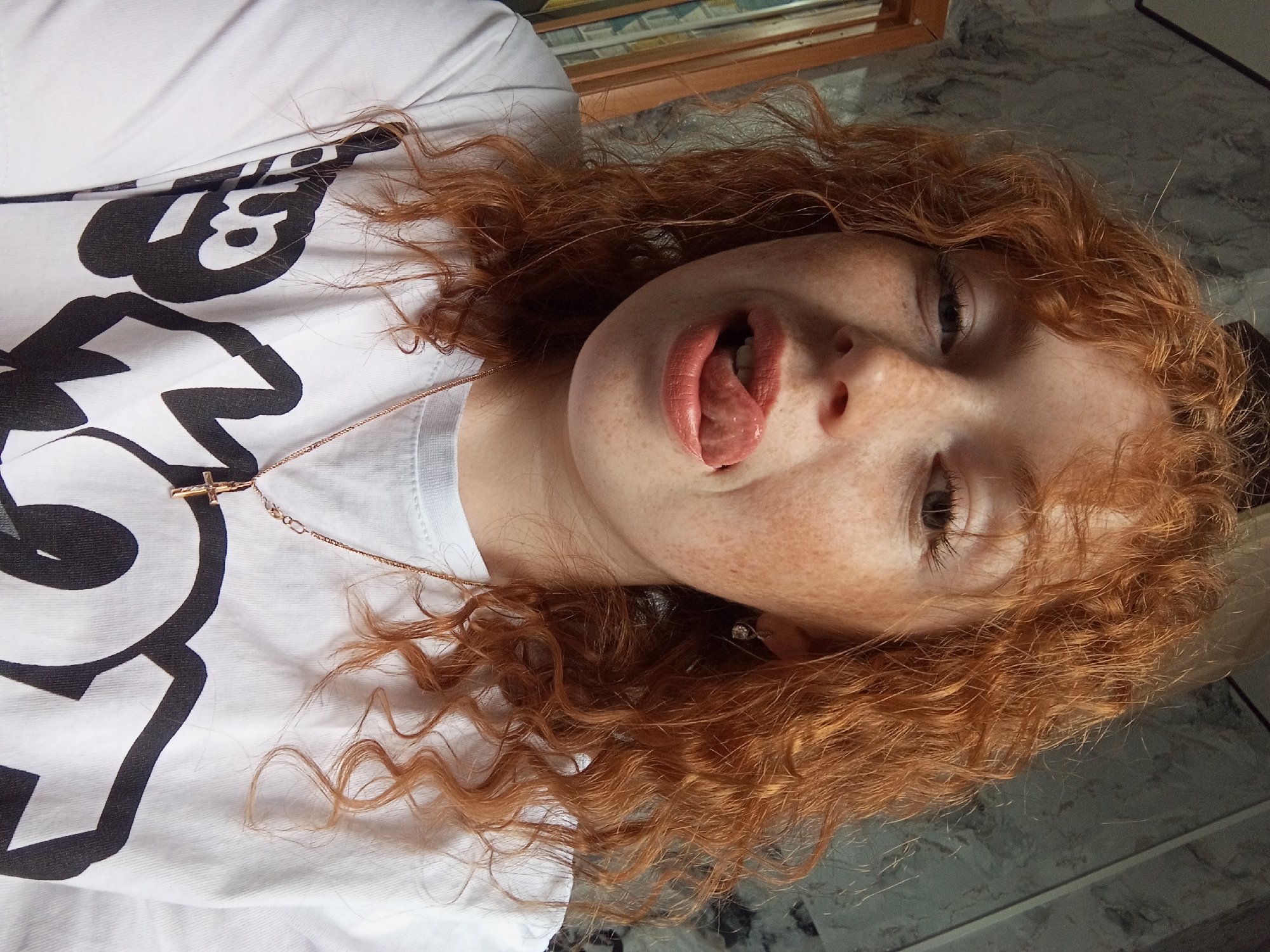 A young, fair-skinned person with red, curly hair is lying on a surface resembling a table or countertop with a marbled pattern. The person is wearing a white T-shirt with a large, stylized black and white graphic, along with a gold necklace featuring a small cross pendant. Their mouth is open, and the tongue is slightly protruding. The background is blurry, but there is a wooden frame visible behind the person's head. There is no indication as to the location of the image.