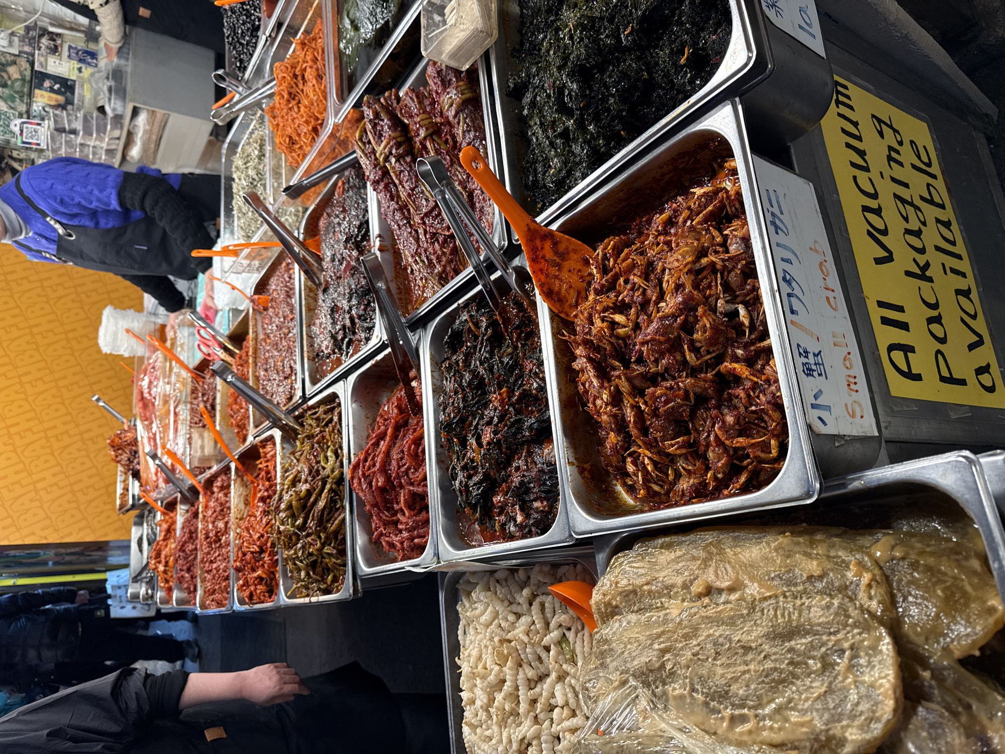 This image depicts a bustling food stall within a traditional market in Seoul, South Korea, likely Gwangjang Market or a similar establishment. The foreground is dominated by multiple rows of stainless steel rectangular trays filled with a wide variety of Korean banchan (side dishes) and specialty food items. Items visible include various forms of red kimchi, marinated green chili peppers, dark pickled greens, fermented small crabs (labeled "Small Crab" in English and Japanese), dried small fish, and a tray of light-colored, textured dried snacks. Numerous metal tongs and orange plastic scoops are present in the trays for serving. On the right side of the frame, a vendor wearing a blue cable-knit sweater, dark apron, and black arm warmers stands behind the counter. Another individual's arm and apron are partially visible on the far right. A yellow sign at the lower right reads, "ALL Vacuum Packaging available." The background shows blurred figures of people walking, and a solid orange wall with an illegible repeating pattern.