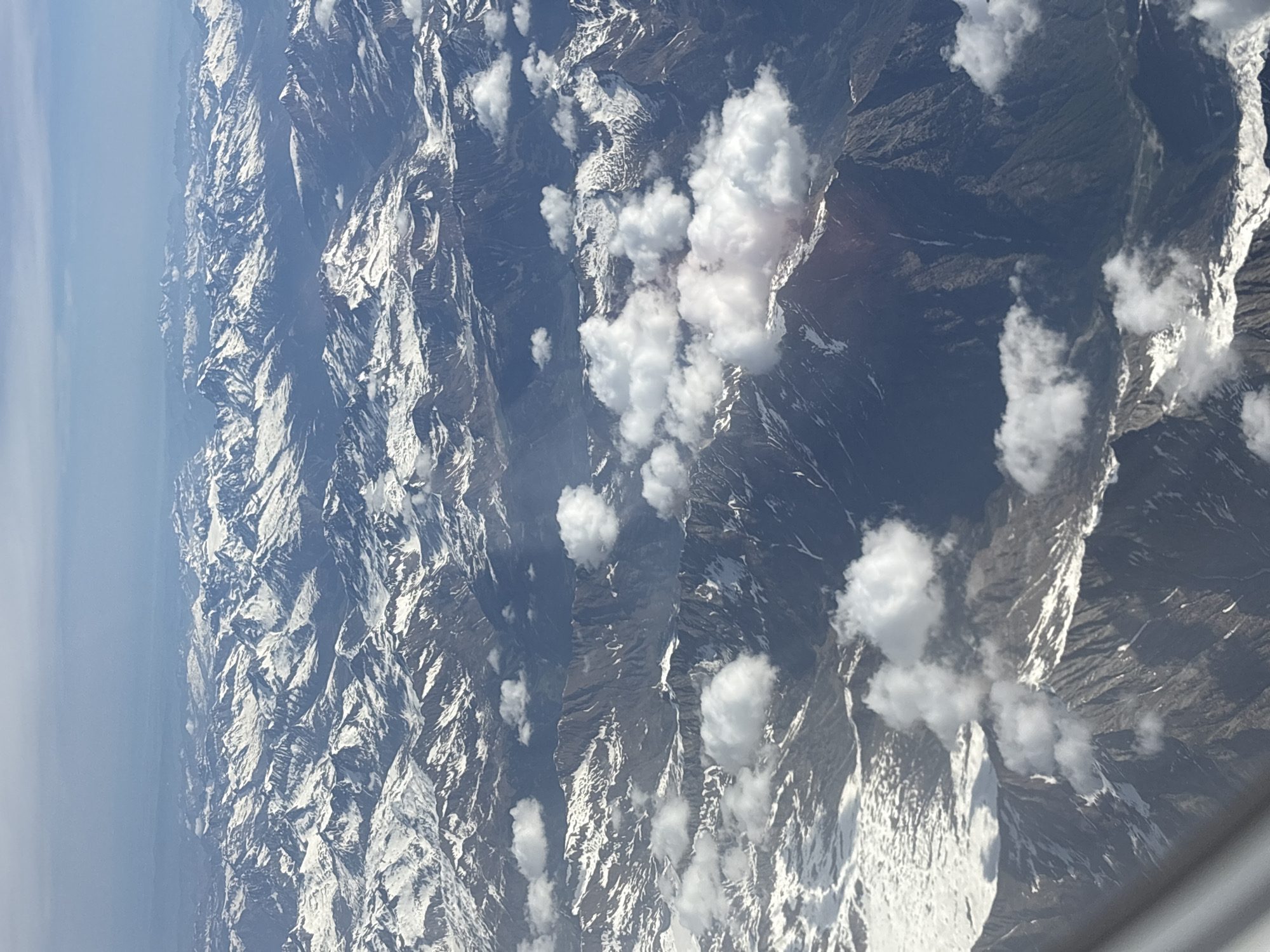The image shows a mountainous landscape covered in snow, interspersed with clouds. The mountain ranges appear rugged and steep. No people are visible. The context lacks specific elements to pinpoint its location in Milan, Italy. The image is shot from an elevated perspective, possibly from an airplane window.