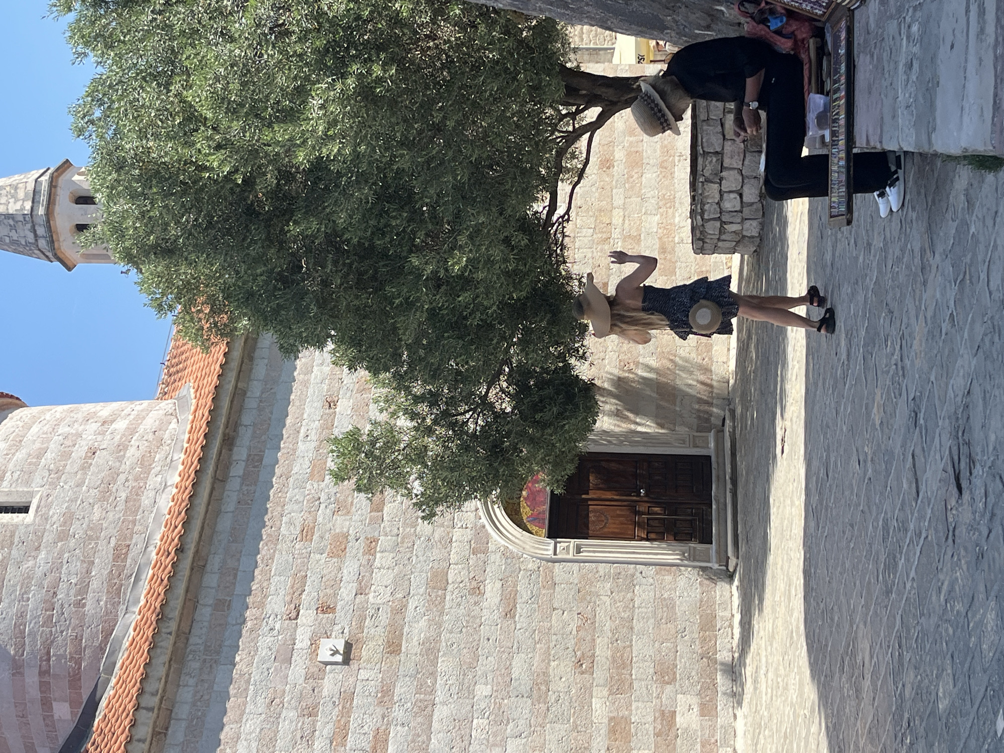 The image depicts a view of the courtyard outside the Church of St. John in Budva, Montenegro. A person wearing a straw hat and a dark patterned dress is standing with their back to the camera, facing a large tree and extending a hand. To the right, an elderly individual with a straw hat and a beard is seated on a bench, engaged in an activity that appears to be selling or arranging small items from a display in front of them. The architectural elements of the church, constructed from stone blocks and featuring a terracotta tiled roof, are visible in the background, along with a distinct tower. The ground is paved with stone. The scene is bathed in sunlight, casting shadows.