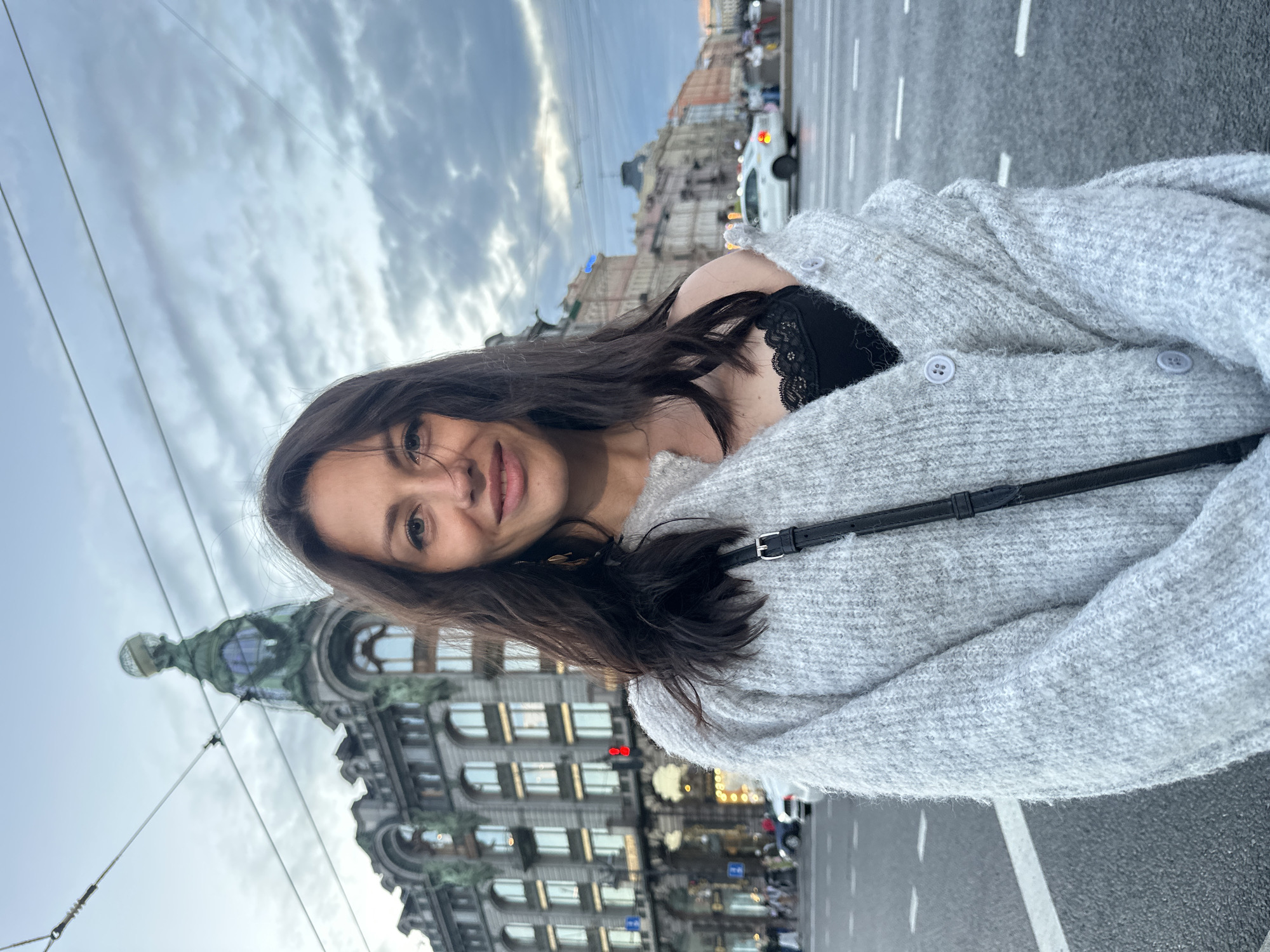 A medium shot captures a young woman standing on a city street, likely in Saint Petersburg, Russia, given the architectural style of the buildings. She is looking directly at the camera with a slight smile, her dark hair falling loosely around her shoulders. She is wearing a light gray, oversized cardigan with large buttons over a black lace-trimmed camisole. A black strap, likely from a purse, is visible across her chest.
Behind her, an ornate, multi-story building with arched windows and a distinctive green, glass-domed spire dominates the background. The building appears to be historic, possibly a landmark. The street in front of the building is paved and has lane markings. Several vehicles are visible on the street, including a white car with its taillights illuminated. Overhead, power lines crisscross the sky. The sky itself is overcast with dramatic clouds, suggesting it may be dusk or an approaching storm. The overall atmosphere is urban and slightly moody.
