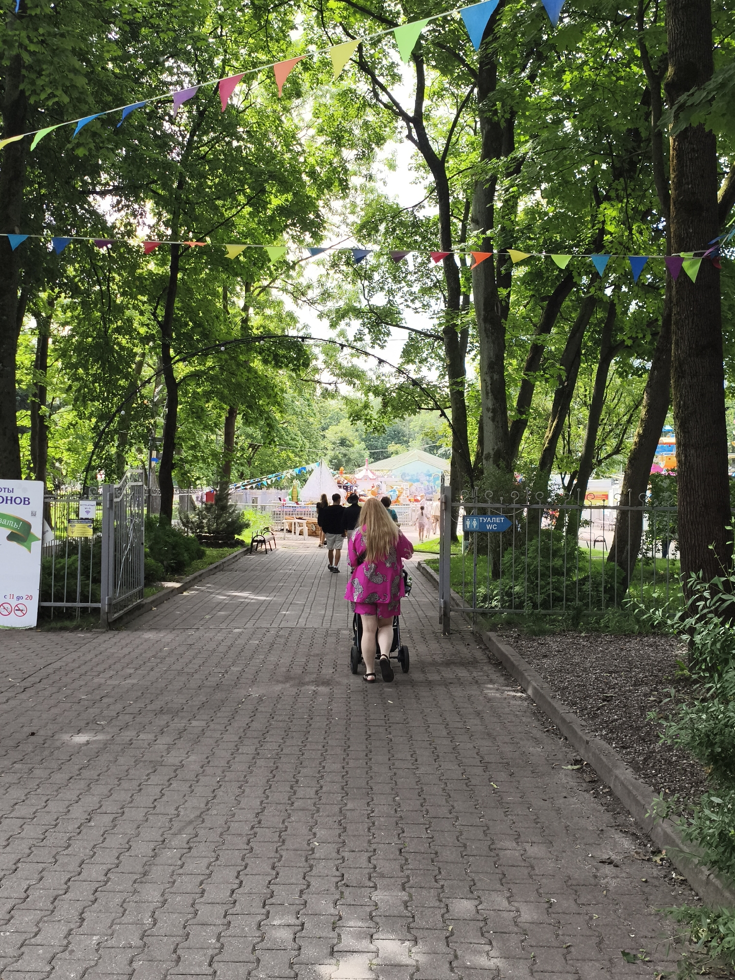 In a paved park pathway lined with trees and decorated with colorful triangular flags, a person wearing a bright pink dress pushes a stroller. The pathway is flanked by metal fences. To the left, a gray metal gate is partially open, revealing a sign with text and graphics. Further down the path, other individuals can be seen walking, and in the background, there are glimpses of amusement park rides, including a carousel and a tent. A blue sign indicating "WC" (restroom) is visible on the right fence. The overall scene suggests a public park or amusement area on a bright day. The presence of Russian text on the signs and the context of amusement parks suggest a location within Russia, possibly Kaliningrad, Russia, given the prompt's requirement.
