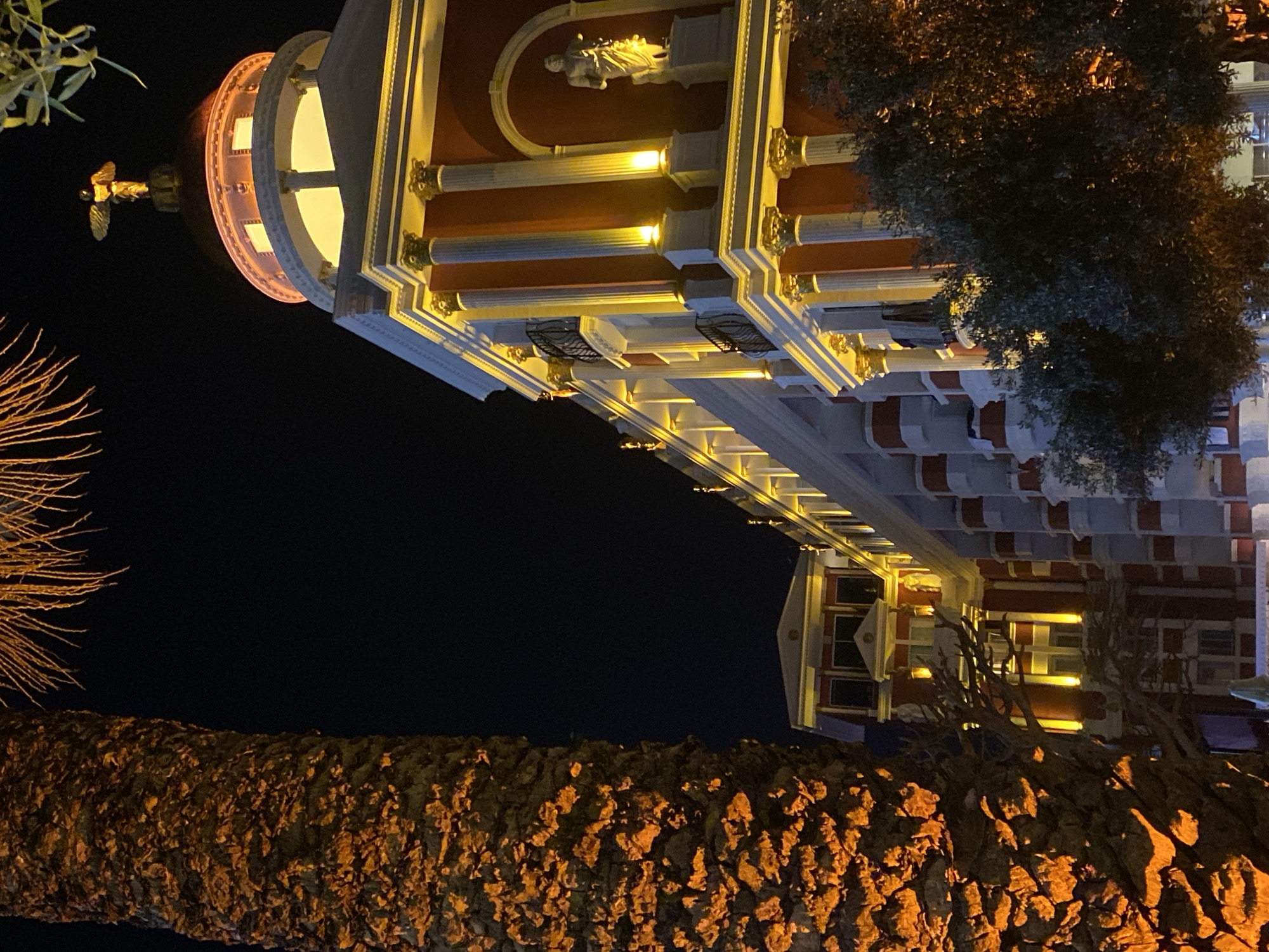 The image depicts the Alanya Municipality Building at night. The architecture features a dome with a golden angel statue on top, white columns with gold accents, red and white walls, and illuminated windows. In the foreground is a tree trunk illuminated by orange lights, with palm fronds visible. The background is a dark night sky.