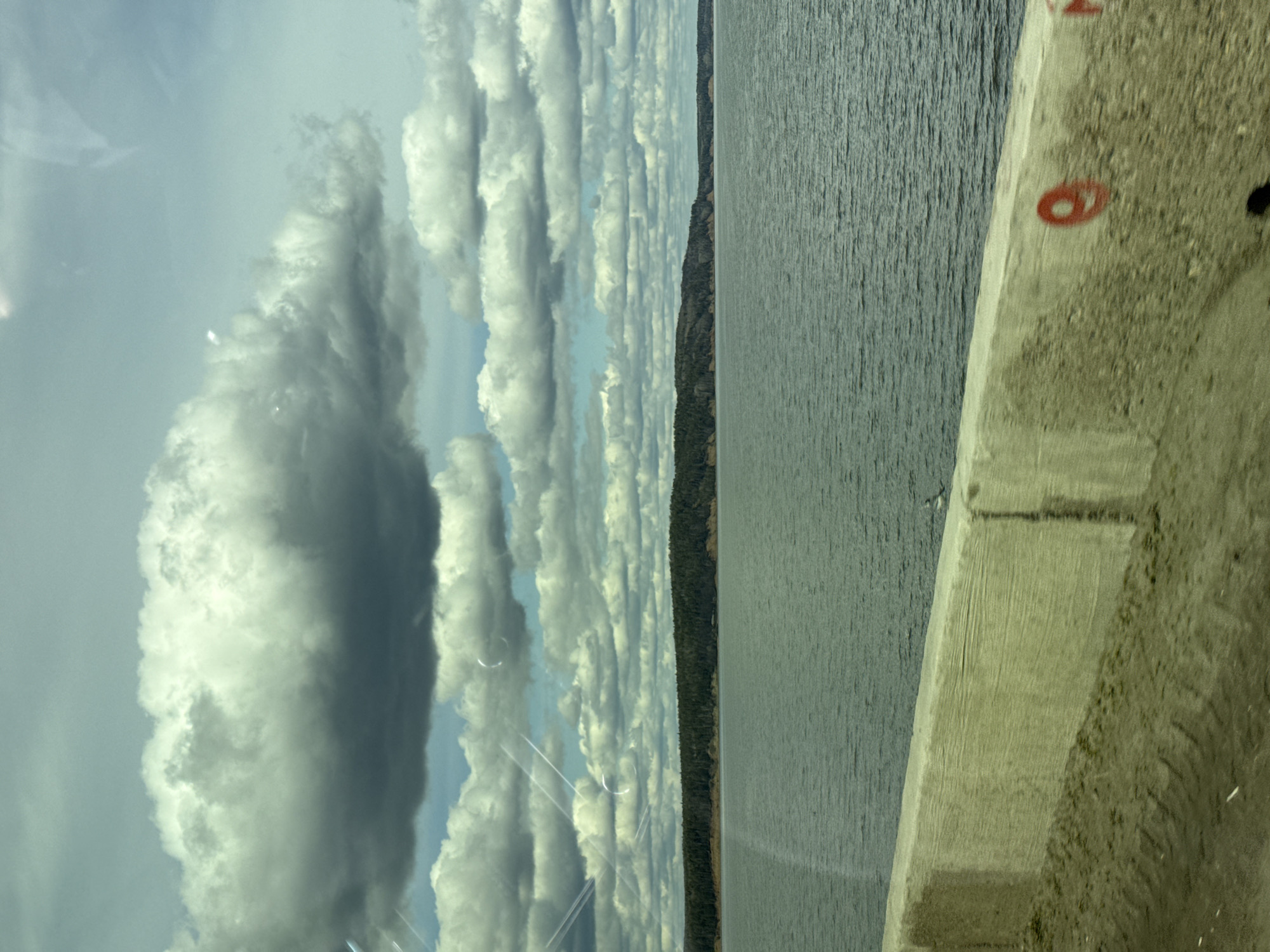 The image, taken in Chaikovsky, Russia, depicts a vast natural landscape from what appears to be a bridge or a high vantage point, possibly through a window given the faint reflections. The scene is dominated by a wide expanse of water on the right, likely a large river or reservoir, and a cloudy sky on the left.
The sky is filled with numerous cumulus and stratocumulus clouds, varying in brightness from brilliant white to darker grey, suggesting depth and differing thickness. Patches of lighter blue sky are visible between the clouds. Subtle, circular and linear reflections or distortions are present in the sky, consistent with viewing through glass.
Below the sky, a distant, dark green forested shoreline stretches horizontally across the middle of the frame, acting as a thin separator between the water and the clouds. The water itself appears calm with a textured surface, reflecting the muted light of the sky.
In the immediate foreground, on the right side of the image, a concrete barrier or railing is visible. It is light beige-grey, rough-textured, and rises diagonally from the bottom right corner towards the center. A distinctive red number "6" is painted on this concrete surface. Further back on the concrete, another partial red marking resembles a circle or another "6."
The lighting suggests it is daytime, possibly mid-day, with even illumination and no strong shadows. No people or animals are visible in the scene.
