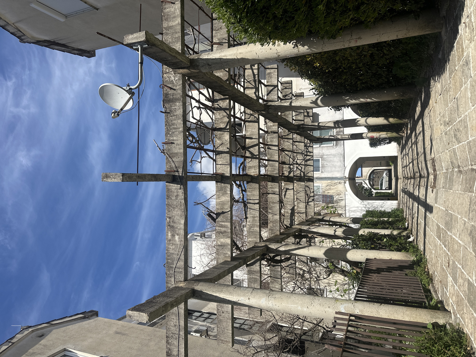 The image presents a pergola structure in Budva, Montenegro, characterized by reinforced concrete posts and beams supporting a metal grid, likely intended for climbing plants. A satellite dish is mounted on one of the posts. The structure frames a pathway paved with rectangular stones, leading towards an arched opening in a white building. The surrounding environment includes vegetation, buildings with windows, and a clear blue sky with thin clouds. The scene appears deserted.
