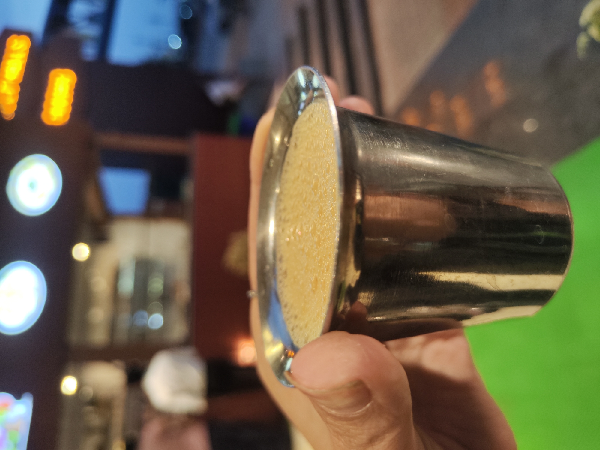A person's hand holds a stainless steel cup filled with a frothy, light-brown liquid. The background is blurred, showing lights, buildings, and a green surface. The location is Mudugurki, India.