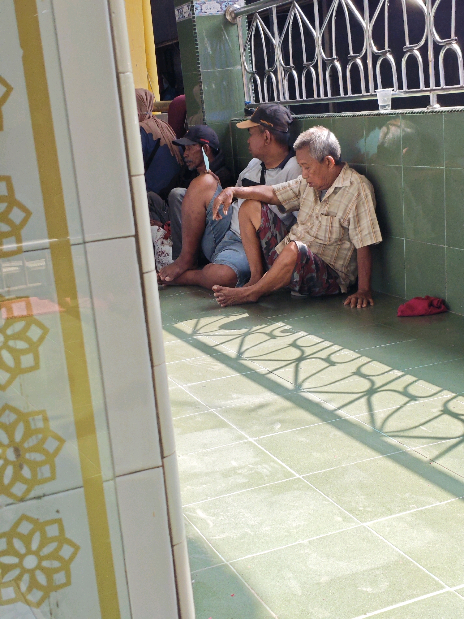 The photograph shows four individuals sitting on the floor against a tiled wall in what appears to be a public area or outdoor waiting space, possibly in Teddaopu, Indonesia. The floor is tiled with large square green tiles and a portion of a white tiled wall with yellow geometric accents is visible on the left. Sunlight casts shadows of an ornamental railing onto the floor.
Three men are clearly visible, and part of a fourth person's head and shoulder is seen behind them. The man in the foreground, on the right, is an older man wearing a checkered short-sleeved shirt and patterned shorts. He is sitting with his legs extended and his left hand resting on the floor. Next to him, to his left, is a younger man in a t-shirt and denim shorts, also seated with his legs bent. Another man, wearing a dark baseball cap and a t-shirt, is seated to the left of the younger man, with a light-colored mask hanging from his face. Behind them, a person wearing a brown headscarf and a dark blue top is partially visible. A red cloth is on the floor to the right of the seated men, and a plastic cup is on a ledge behind them. The wall behind the people is tiled with green squares, and a metal railing with an intricate design is mounted above it. The overall impression is a scene of people resting or waiting in a shaded area, illuminated by sunlight.
