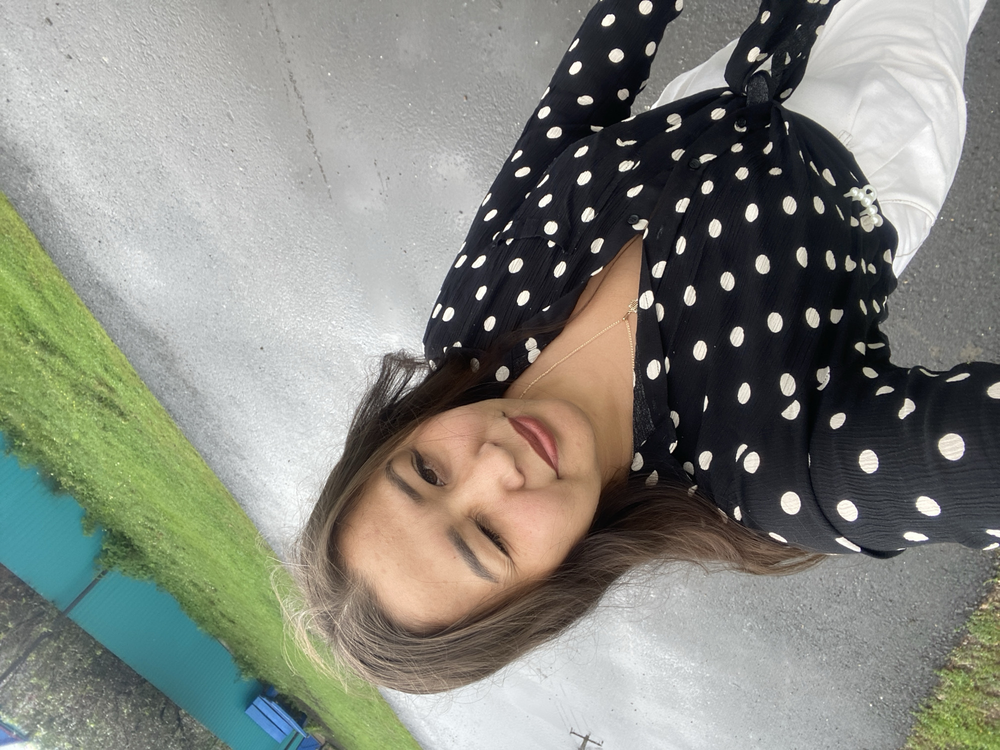 A young woman is taking a selfie outdoors in Maloarslangulovo, Russia. She is winking at the camera and wearing a black polka dot blouse tied at the waist and white pants. Behind her, a green fence and grassy area are visible, along with a paved surface that appears wet, suggesting it may have recently rained. A blue bench is also present in the background. The lighting suggests it is daytime.