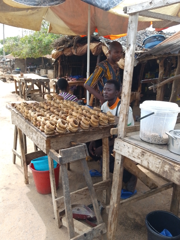 The image depicts an outdoor market stall in Igbo Ijaiye, Nigeria. In the foreground, a rustic wooden table is laden with numerous light-brown, ring-shaped food items, stacked in multiple rows. To the right, a second wooden table holds a clear plastic bucket containing light-colored grains or legumes, and a small metal pot.
Three individuals are present. A bald adult male, wearing a patterned blue, yellow, and brown tunic, stands behind the main display table, facing slightly left. Below and to his right, a second adult male with short dark hair is seated or crouching, wearing a light-colored t-shirt with orange trim, looking to the right. A third individual, seen from behind, wearing a striped shirt, is partially visible in the middle ground, further back between the two men.
The scene is shaded by an overhead, light-colored umbrella canopy. The ground consists of compacted dirt. In the background, several rudimentary market structures constructed from wooden poles and corrugated metal sheets are visible, with foliage appearing beyond them. Various objects are on the ground, including a red bucket containing a light blue basin, a dark blue bucket, and several pairs of blue flip-flops. A small red and black object lies beneath the main display table.