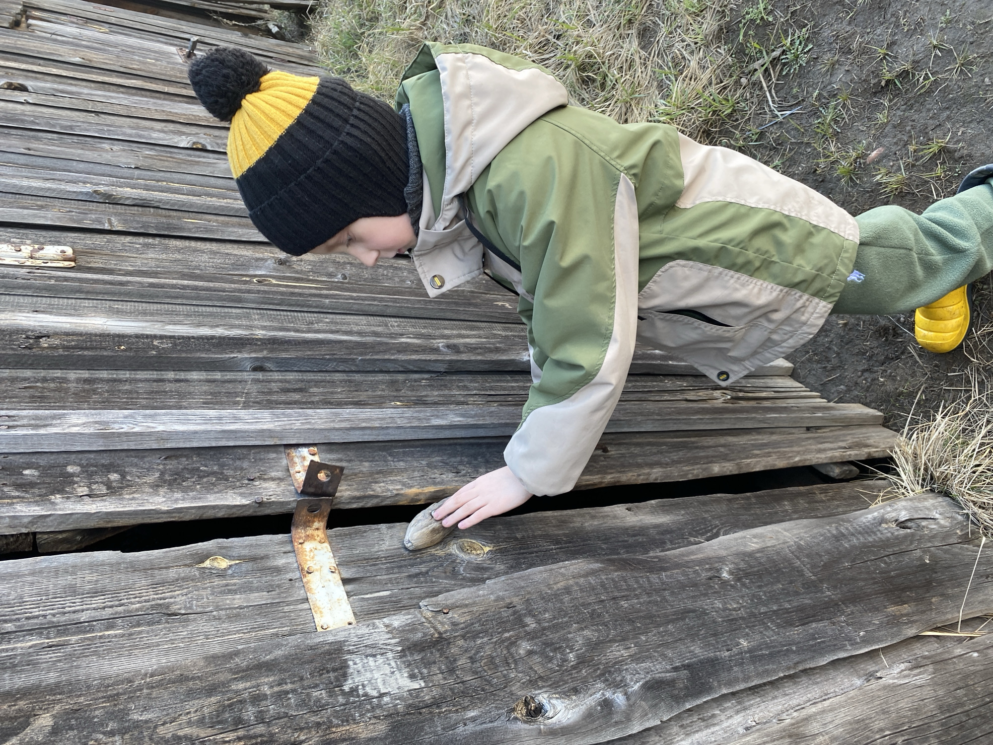 A young person wearing a black and yellow knit hat, a green and beige jacket, and green pants with yellow boots is leaning over a weathered, gray wooden structure, possibly a bridge or deck. The person's hand is resting on the wood. The structure has gaps between the planks, and some rusty metal brackets are visible. The background includes some dry grass and dirt. The image was taken in Noviy, Russia.
