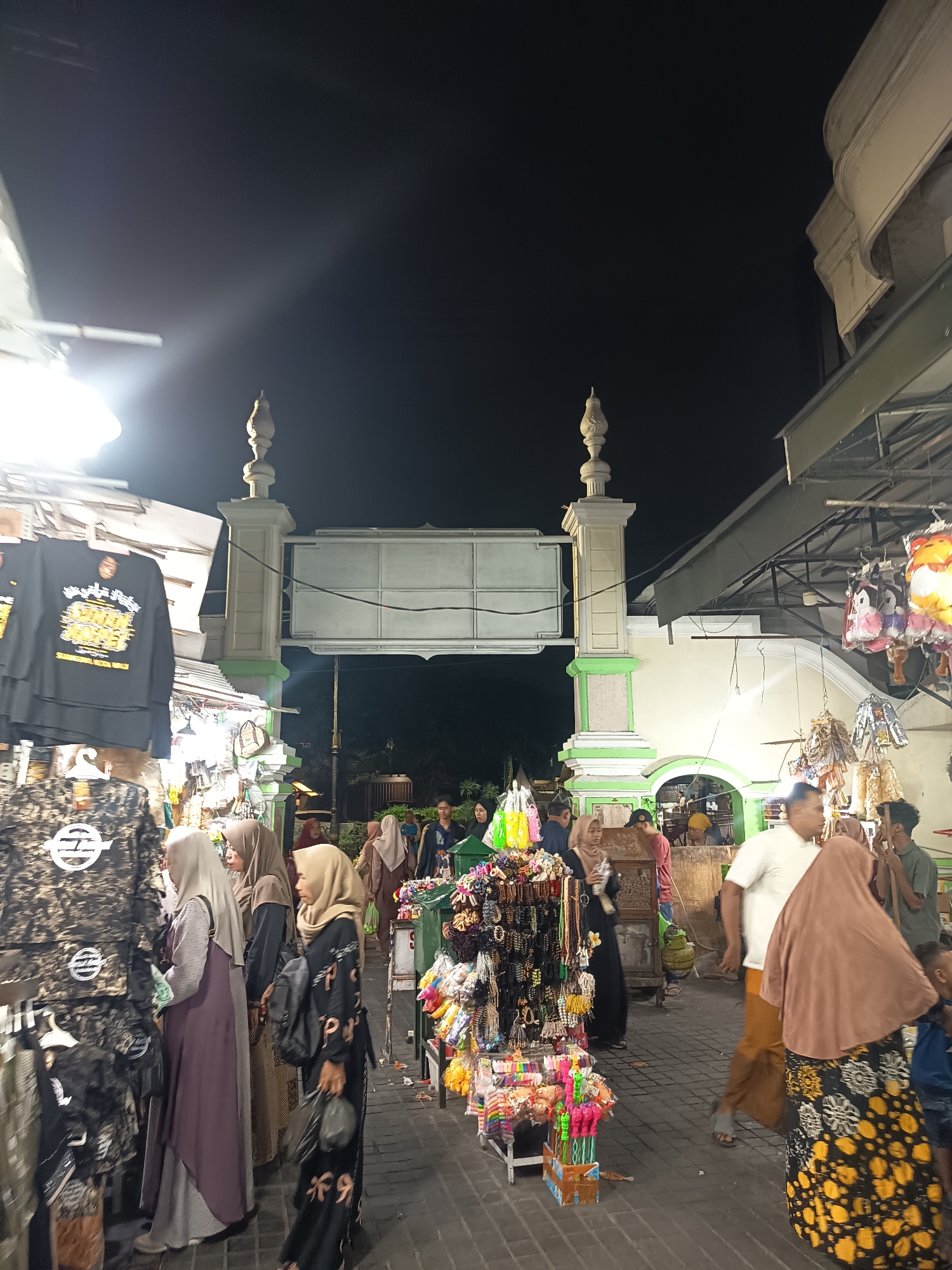This is a night-time street scene in front of the Ampel Mosque in Surabaya, Indonesia. The image captures a bustling atmosphere with several people browsing market stalls that sell various items, including clothing, accessories, and toys. In the foreground, people are seen examining the merchandise. To the left, racks of t-shirts with intricate designs are displayed. Further back, a group of individuals are walking through the crowded pathway. The Ampel Mosque's characteristic gateway, adorned with green and white pillars and arches, stands prominently in the background, illuminated by the surrounding lights, creating a vibrant and lively ambiance.