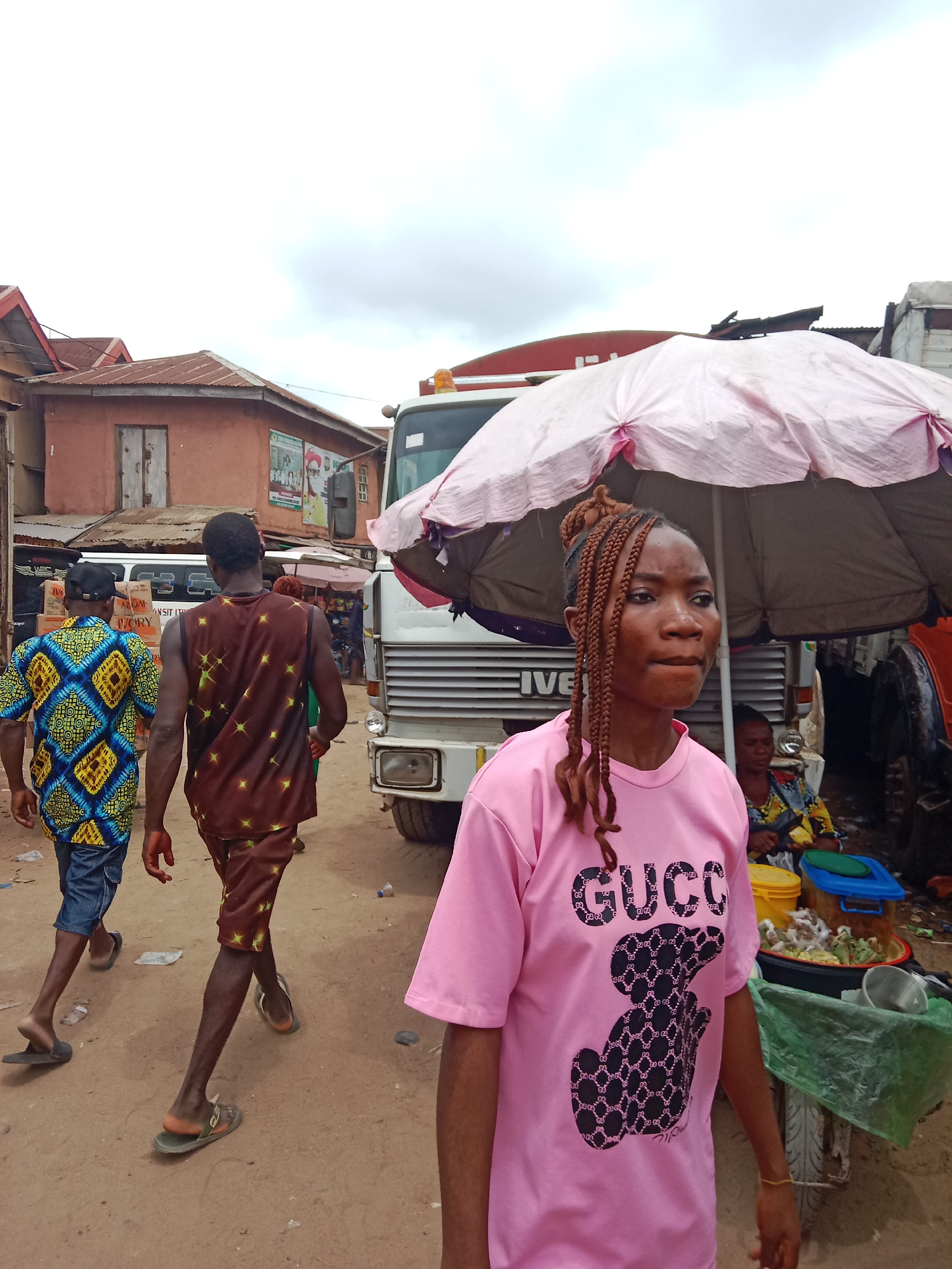 The image depicts a street scene in Onitsha, Nigeria. A young woman wearing a pink t-shirt with a black "Gucci" teddy bear graphic is prominently featured in the foreground, looking off to the right. Behind her is a white Iveco truck parked next to a stall covered by a faded pink umbrella. An older woman sits at the stall, surrounded by food items in containers. Further back, two men walk away from the camera, one wearing a blue and yellow patterned shirt and jeans, the other in a brown patterned outfit. The buildings are simple and painted in reddish-brown and other muted colors. The ground is dirt. The sky is overcast.