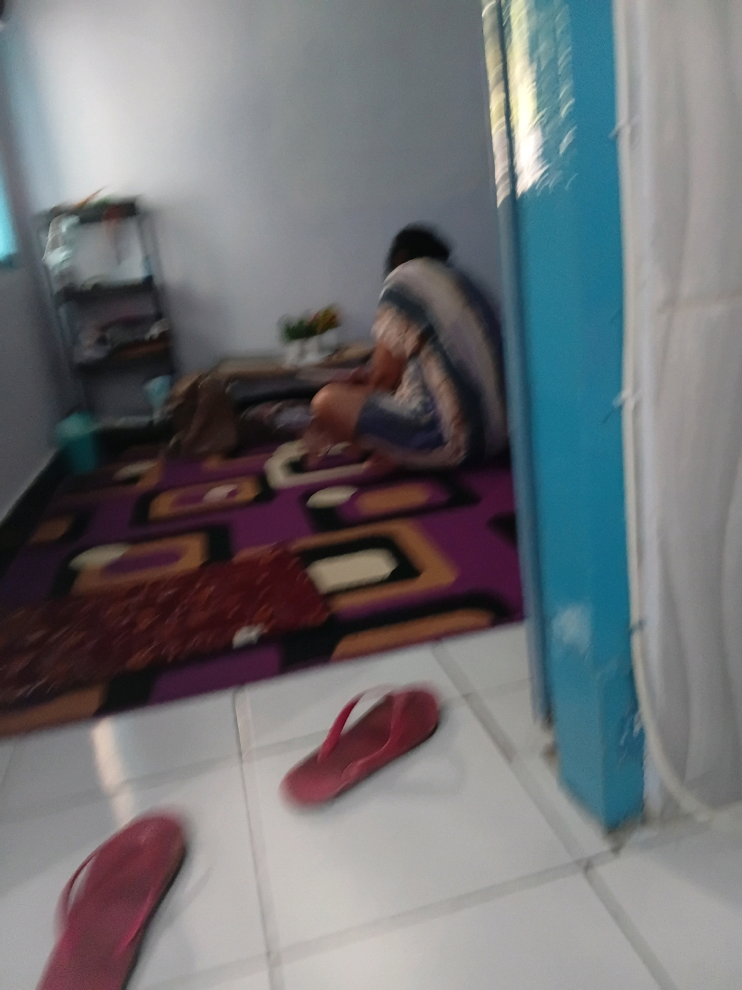 The image depicts an indoor scene, likely a residential room, located in Tanggul Wetan, Indonesia. In the background, a person is seen sitting on a carpeted floor, facing away from the camera. The person is wearing a striped garment. In the foreground, two red flip-flops are visible on a tiled floor. A shelf unit is present on the left side of the image. The overall lighting suggests it is daytime. The image is blurry, making precise identification of all details challenging.