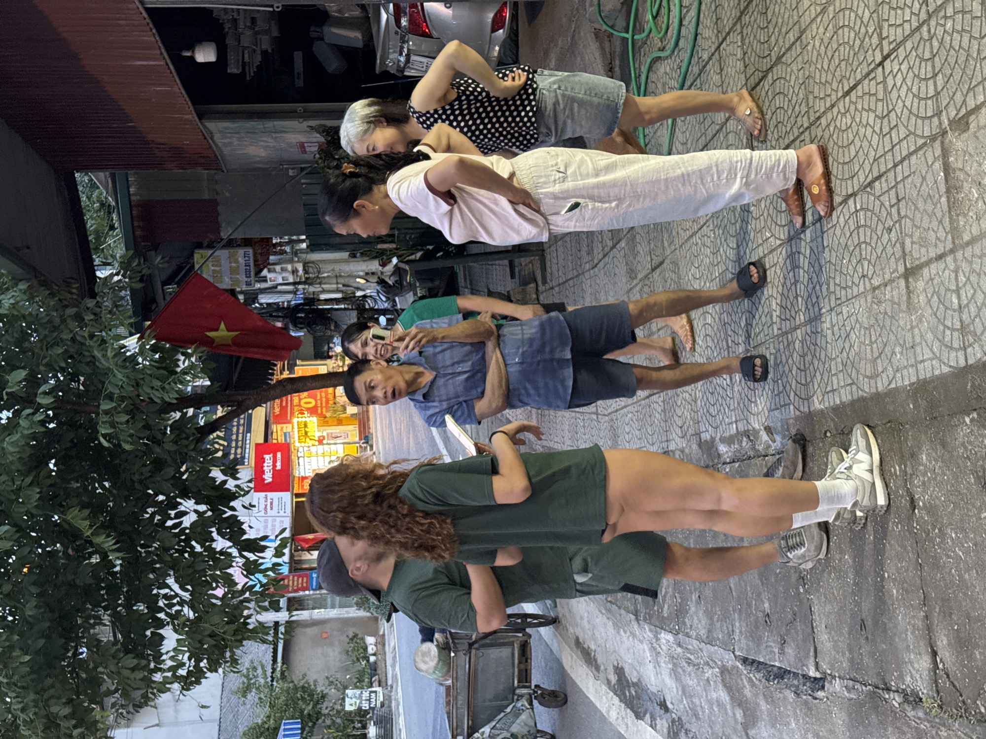 The image depicts a vibrant street scene in Vietnam, likely during late afternoon or early evening. The setting is an outdoor urban or semi-urban environment, with a patterned sidewalk, buildings lining the street, and a large leafy tree on the left.
A diverse group of people is present, engaged in various activities. In the foreground, a couple shares an intimate moment; a woman with long, curly brown hair in a green t-shirt and light shorts is embracing a man in a dark cap, green t-shirt, and green shorts. Behind them, a street vendor's cart with a green bucket is visible.
Further back, a group of three individuals appears to be interacting with a camera. A man in a blue denim shirt and dark shorts holds a phone, looking towards the viewer, seemingly posing for a photo alongside a partially obscured woman. Another man in a green t-shirt is also holding a phone, potentially taking a photo or video.
In the background, two women stand casually; one with short gray hair wears a polka dot top and denim shorts, while the other with dark hair wears a white t-shirt and white pants. A green garden hose lies on the sidewalk near them.
Key location indicators include a prominent Vietnamese flag (red with a yellow star) hanging on the left, and various storefronts with signs. Visible text includes "TÓC NAM" (Men's Hair), "CƯỜNG XUÂN MOBLE", "viettel telecom" (appearing twice), "0%", and a phone number "DT: 6332.811.771". The atmosphere is casual and social, consistent with a street gathering in Vietnam. While the specific iconic landscapes of Mù Cang Chải are not visible, the scene reflects daily life in a Vietnamese town or city.