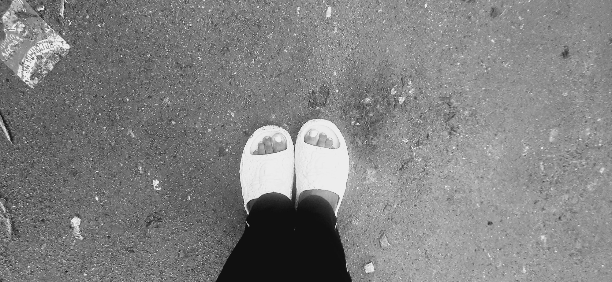 This monochrome, top-down photograph captures a person's lower legs and feet, adorned in white slide sandals, standing on a textured ground surface. The sandals feature an embossed pattern. The visible parts of the feet show the toes, with no discernable polish on the toenails. The person is wearing dark pants or leggings.
The ground appears to be a rough, granular surface, likely asphalt or concrete, consistent with an outdoor setting in Jos, Nigeria. Small specks and debris are scattered across the ground, indicating a public or less-maintained area. In the upper left corner, there is crumpled litter, possibly paper or plastic packaging. Faint, partially readable text on this debris includes the word "Brands" and what seems to be "REC" or "RPO C".
The image is shot with even, diffused lighting, preventing clear shadows and making the time of day undeterminable. The subject appears to be stationary, with the focus on the immediate ground and footwear. The overall scene suggests an ordinary moment in an urban or semi-urban environment.