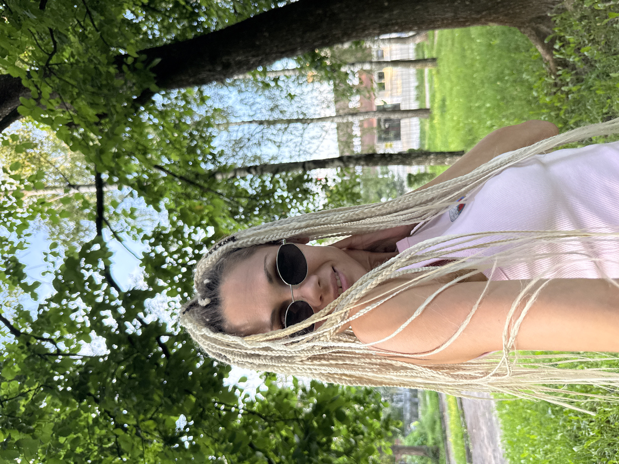 A young woman with blonde braided hair and sunglasses is pictured from the chest up, in front of a blurred background of trees and grass. She is wearing a pink tank top. The location is identified as Rusanovo, Russia. The image appears to be taken outdoors during the daytime, with sunlight filtering through the leaves of the trees. The woman is looking at the camera and has her hand near her neck.