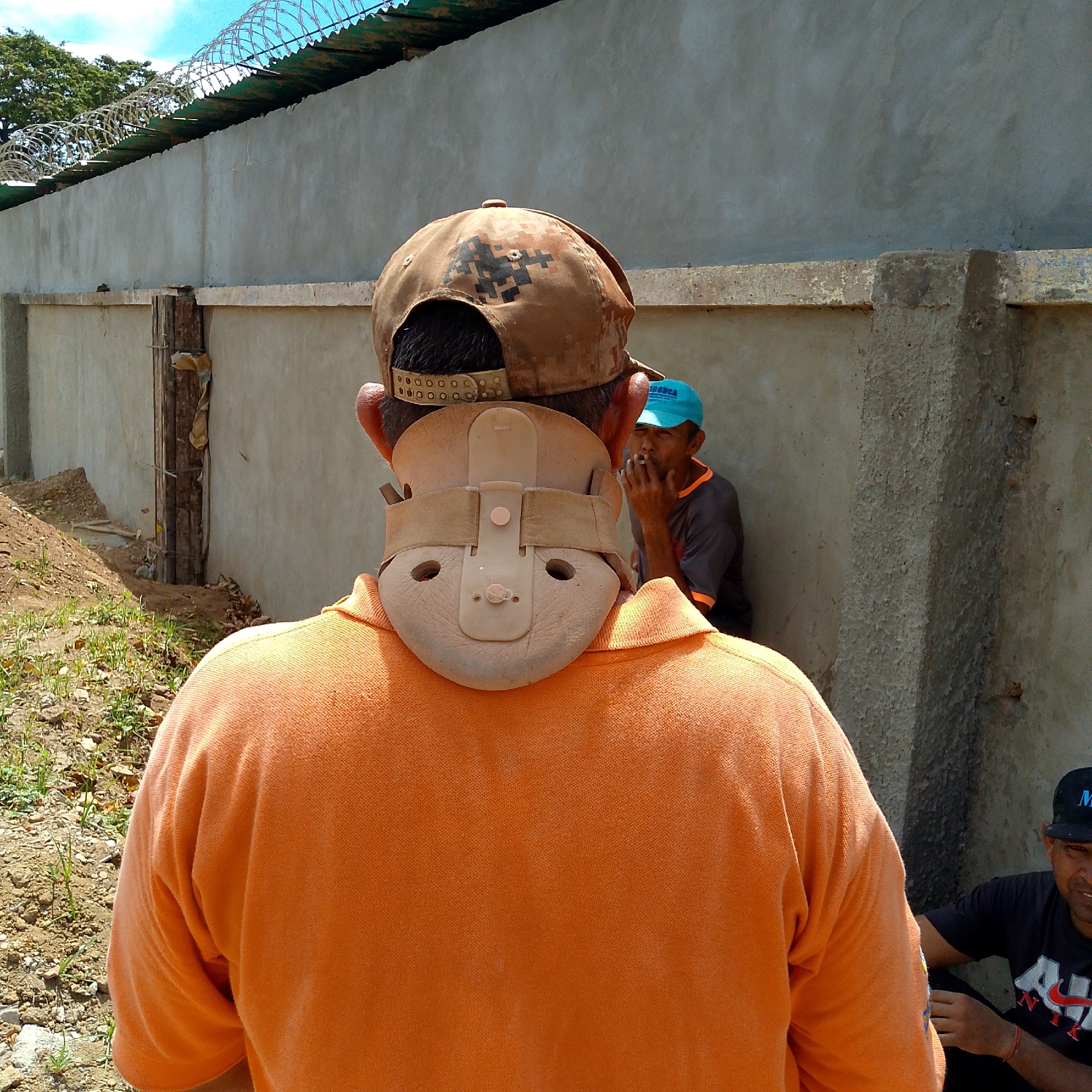 The image depicts the back of a person wearing an orange shirt, a neck brace, and a baseball cap, standing outdoors. Behind the person, two other individuals are visible. One is smoking, wearing a blue cap and a gray shirt. The other is seated, wearing a black cap and a black t-shirt with a red and white logo. A concrete wall with barbed wire on top serves as the background. The location is Altagracia, Venezuela.