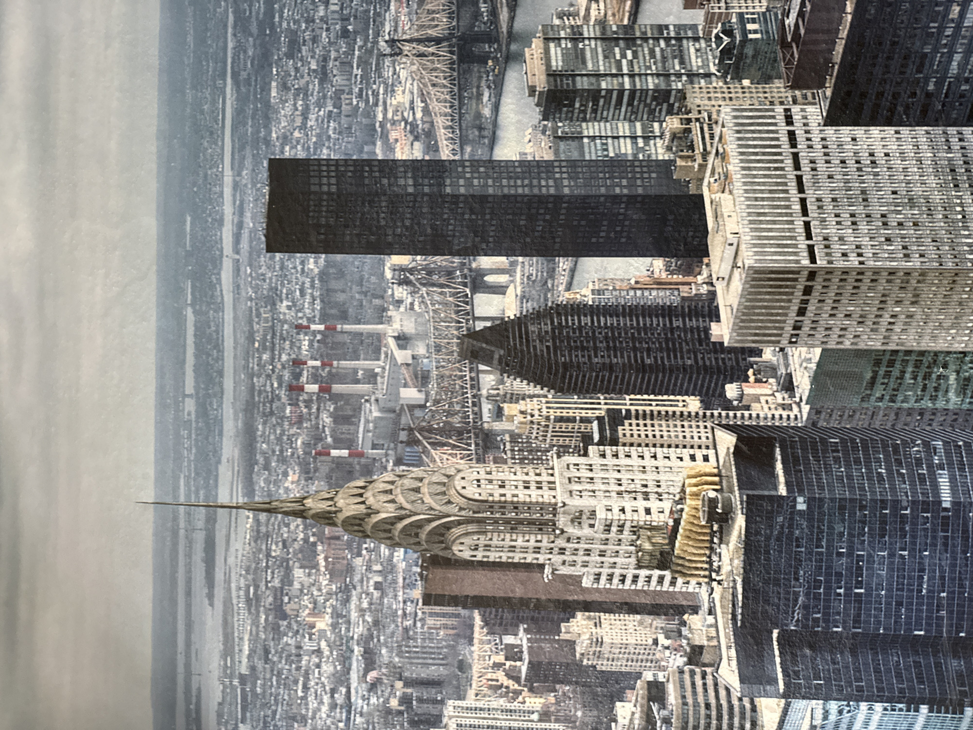 This is a high-angle aerial photograph of the New York City skyline. The image captures a dense urban landscape with numerous skyscrapers of varying architectural styles and heights. Dominating the foreground are several large buildings, including a distinctive dark, rectangular skyscraper and a lighter, patterned building. In the middle ground, the iconic Chrysler Building with its art deco spire is prominently visible, surrounded by other tall structures. Further in the distance, a bridge spans across a body of water, with industrial smokestacks emitting smoke on the far bank. The sky is overcast and hazy, contributing to a muted color palette across the scene. The photograph does not depict Montana, Bulgaria.