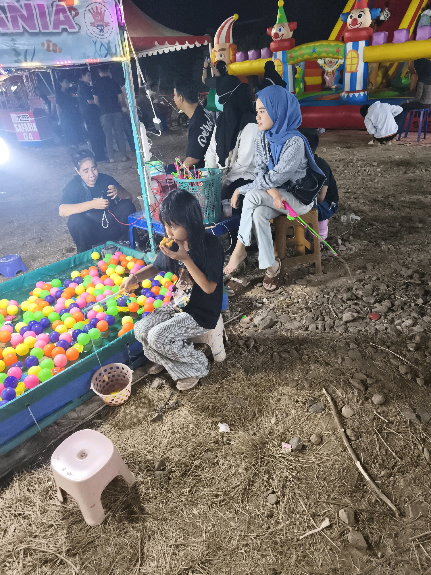 The image depicts an outdoor nighttime scene at a carnival or fairground in Palembang, Indonesia. In the foreground, a young girl with long black hair is crouched by a shallow pool filled with numerous brightly colored balls, including pink, orange, purple, green, and yellow. She is holding a fishing rod with a green string and a pink hook, and she is biting into a piece of food. To her left, an adult woman in a black t-shirt is seated and appears to be holding a beaded necklace. Behind them, several other people are visible, some standing and some seated.
In the background, a large inflatable bouncy castle with clown figures is illuminated. Tents with banners and lights are also present, suggesting a festive atmosphere. The ground is covered in dry grass, dirt, and small rocks. A small pink plastic stool is positioned in the lower-left quadrant of the image. The overall lighting is dim, with artificial lights from the carnival providing illumination.