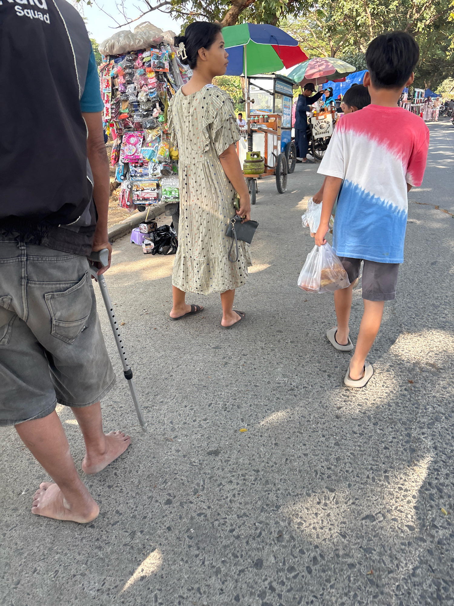 A group of people are walking on a street in Telagamurni, Indonesia. In the foreground, a person is walking with a cane, wearing grey shorts and no shoes. To their right, a woman in a patterned dress and flip-flops is walking. Further ahead, a young boy in a red, white, and blue tie-dye t-shirt and shorts is carrying a plastic bag. In the background, there are several street vendors with colorful umbrellas and carts, selling various items. Trees line the street, and the ground is asphalt. The scene appears to be a busy marketplace.