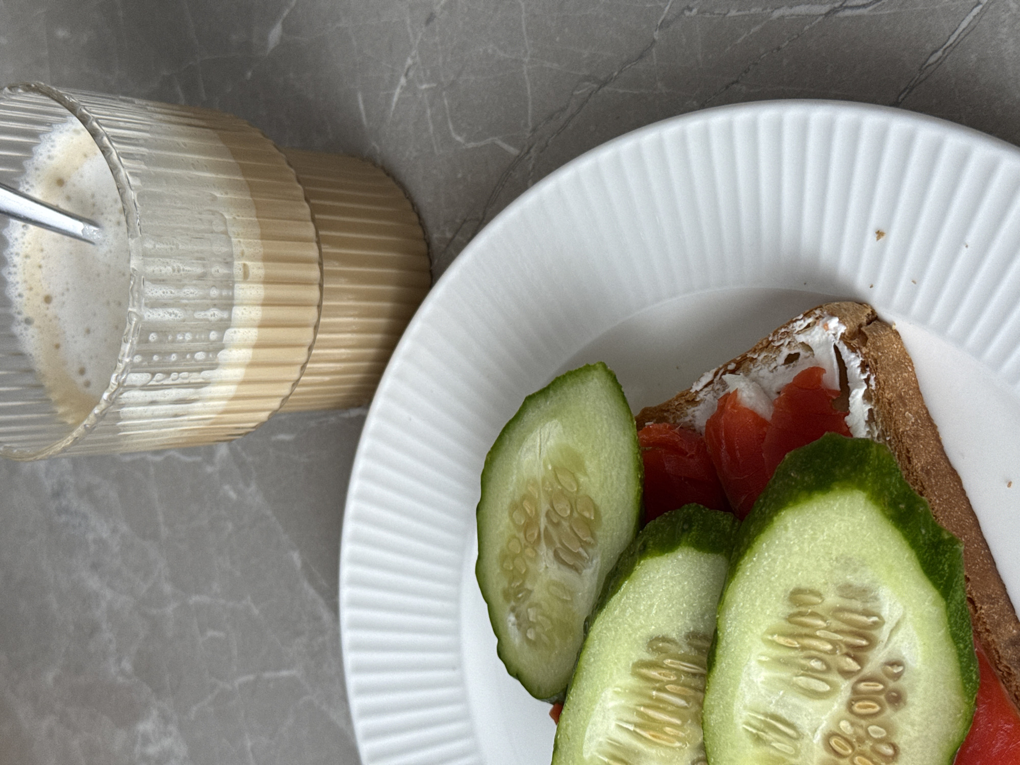 An overhead shot captures a breakfast or brunch spread laid out on a grey, marbled surface. The composition is split diagonally, with a white, ribbed plate occupying the right and bottom portion, and a clear, fluted glass situated in the upper left.
On the white plate, there is a piece of dark bread or toast generously spread with white cream cheese, topped with slices of bright orange-red salmon or cured fish. Alongside the bread, several thick slices of cucumber, still retaining some of their dark green peel, are arranged. The cucumber slices reveal their internal seed structure.
The fluted glass contains a light beige or brown liquid with a substantial layer of white foam or froth on top, suggesting a coffee drink like a latte or cappuccino. A metal spoon is partially submerged in the drink, indicating it might have just been stirred or is about to be.
The overall scene suggests a casual indoor setting, likely a kitchen or dining table, with natural light illuminating the food and drink. No people are visible, and there is no discernible text in the image.