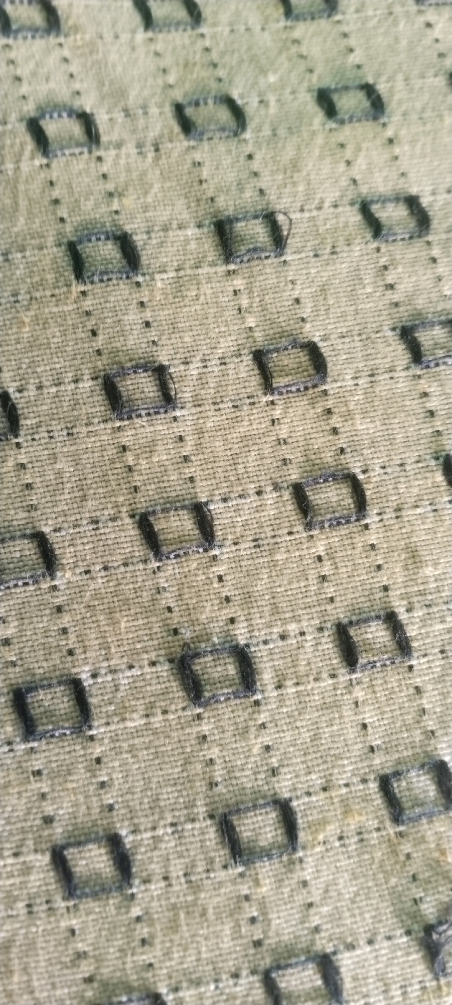 This is a close-up, angled shot of a textured, light green fabric with a repeating pattern of small, dark gray embroidered squares. The squares are arranged in a grid with a dotted line connecting them horizontally and vertically, creating a subtle checkered effect. The fabric appears to be a natural fiber, possibly linen or cotton, with visible weave and some loose threads. The lighting is even, highlighting the texture of the material. This image does not depict a scene in Bhaktarahalli, India; it shows a close-up of fabric.