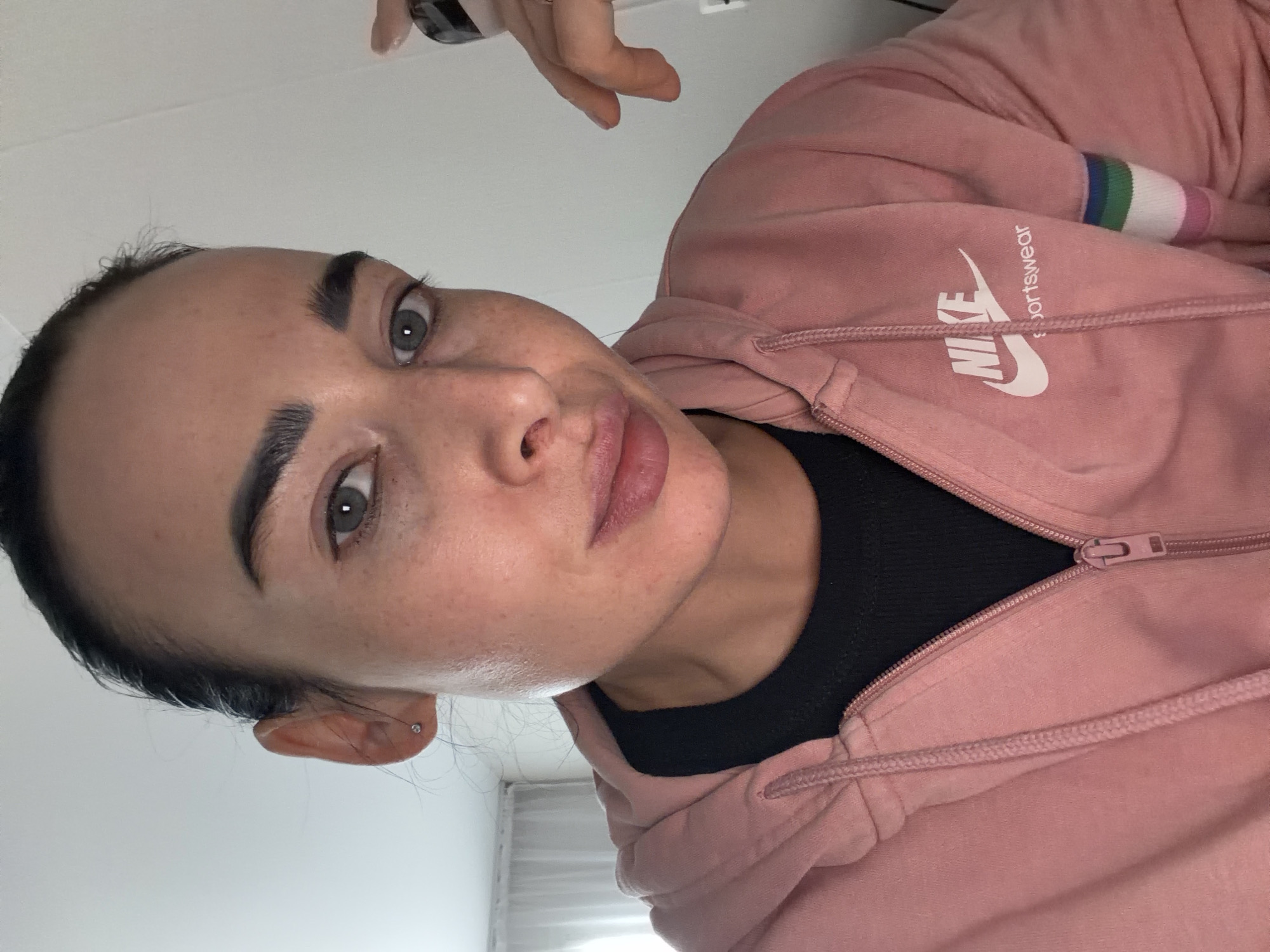 A woman with dark hair and eyebrows is wearing a pink Nike sweatshirt over a black shirt. The sweatshirt has a white Nike logo and the word "Sportswear". The woman is looking at the camera with a slight smile. She has an earring in her left ear. The background is a white wall and a sheer white curtain. There is no indication of the location being Saint Petersburg, Russia.