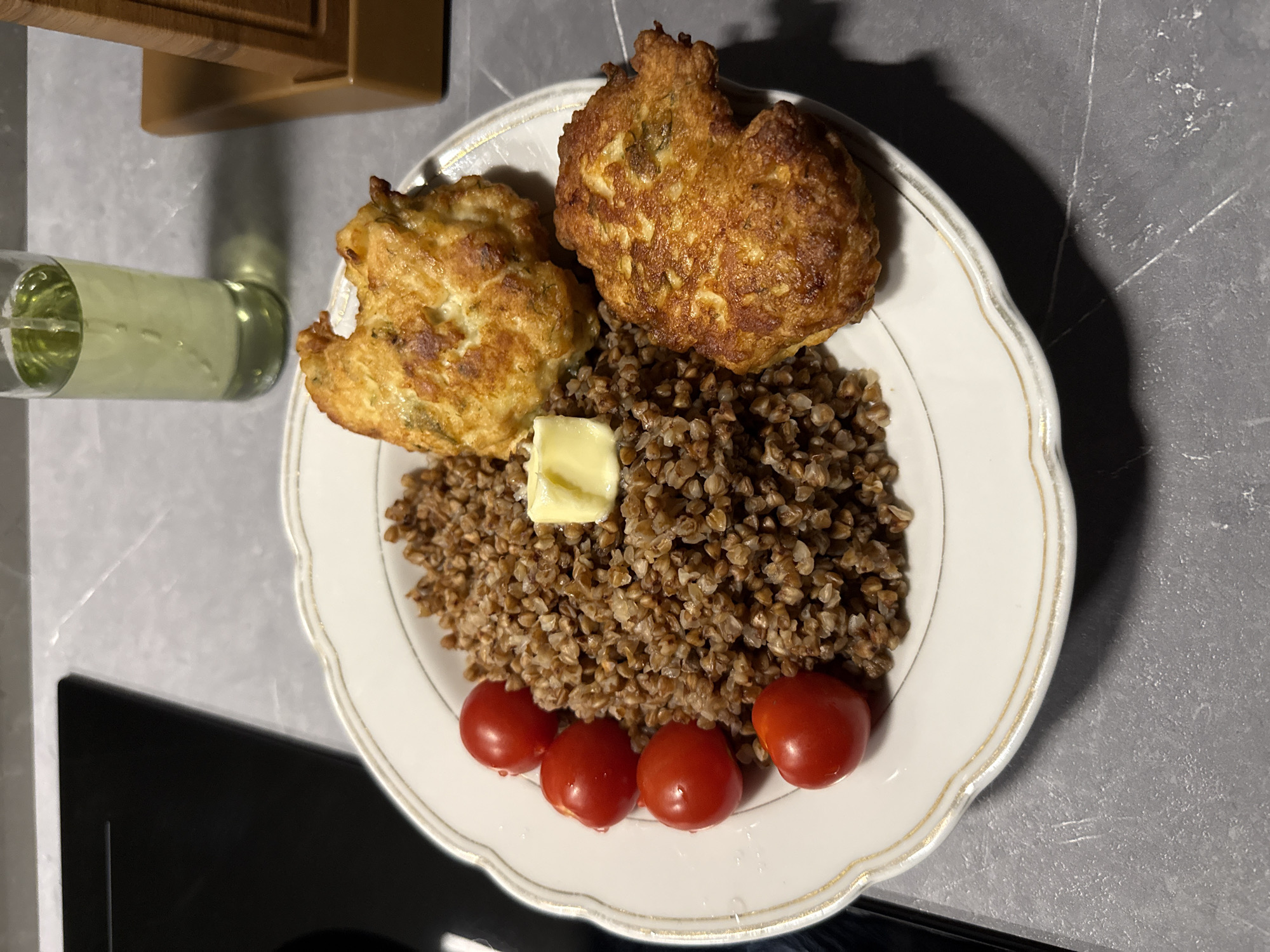 A white ceramic plate with gold trim is filled with food. In the center of the plate is a mound of cooked buckwheat. To the left of the buckwheat are four small, round, red cherry tomatoes. To the right of the buckwheat are two golden-brown, fried patties. A small cube of yellow butter sits atop the buckwheat. In the background, to the left, is a black stovetop. To the right, there is a clear glass with a light-yellow liquid and a wooden object. The surface beneath the plate appears to be a gray countertop.
