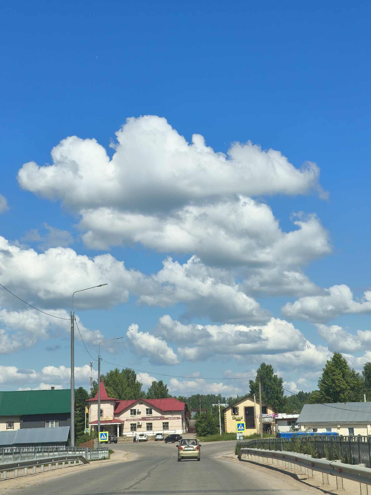 In Kady, Russia, on a clear day, a suburban landscape unfolds under a vibrant blue sky dotted with fluffy white clouds. The scene is captured from a vehicle traveling on a paved road, indicated by the guardrail on the right. In the distance, a cluster of buildings forms a small commercial area.
To the left, a building with a dark green sloped roof and a grey section is visible. Adjacent to it, a building with a red tiled roof and a pointed red turret stands prominently. Signs on this building indicate a store, with Russian text including "ПРОДУКТЫ" (products) and "УНИВЕРСАМ" (supermarket). Several cars are parked in front of these establishments, and a few pedestrians are present.
Further to the right, a smaller yellow building displays a sign with "ZONE" and a yellow pedestrian crossing sign is prominently placed. Another building with a grey sloped roof is situated to the right of this. A cell tower is visible in the background behind the buildings.
The road curves gently to the right as it approaches these buildings. The overall impression is a typical scene of a small Russian town on a sunny day.