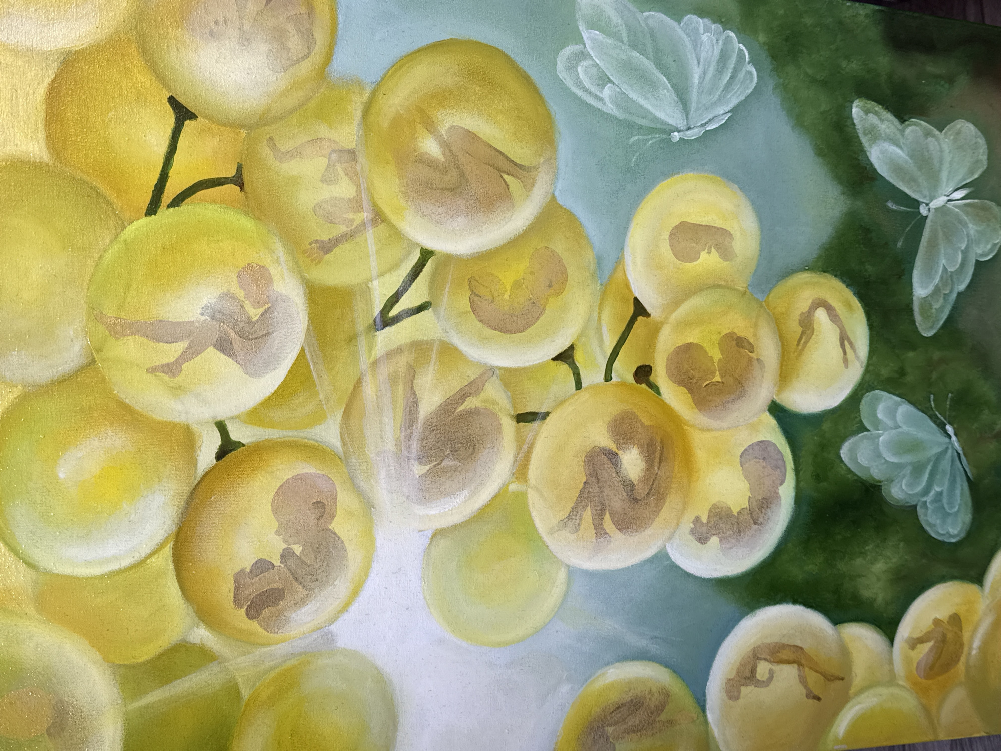 The image depicts a painting featuring a cluster of grapes. Each grape contains a human figure in various stages of development, from fetal positions to seated adults. Two butterflies are present in the background. The location is not evident within the image, and there's no mention of Batumi, Georgia, in the image itself.