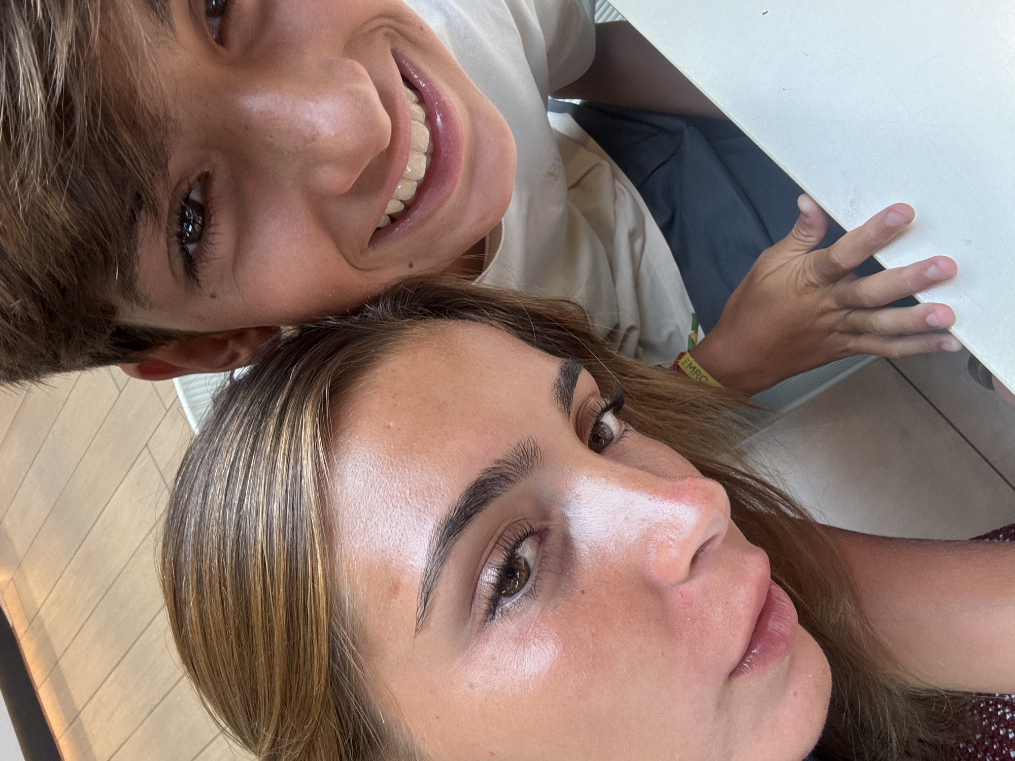 In this close-up, high-angle selfie, two young individuals are captured. The person on the left, a female with long, light brown hair and fair skin, is positioned in the foreground, her face tilted towards the camera and her expression neutral. To her right, a male with short, dark brown hair and a warm smile is visible, his gaze directed at the lens. Both are wearing casual attire. They appear to be seated at a white table. The background suggests an indoor setting with tiled flooring and hints of outdoor elements, possibly a cafe or restaurant, though no specific location in Lisbon, Portugal, can be definitively identified from the visual information provided.