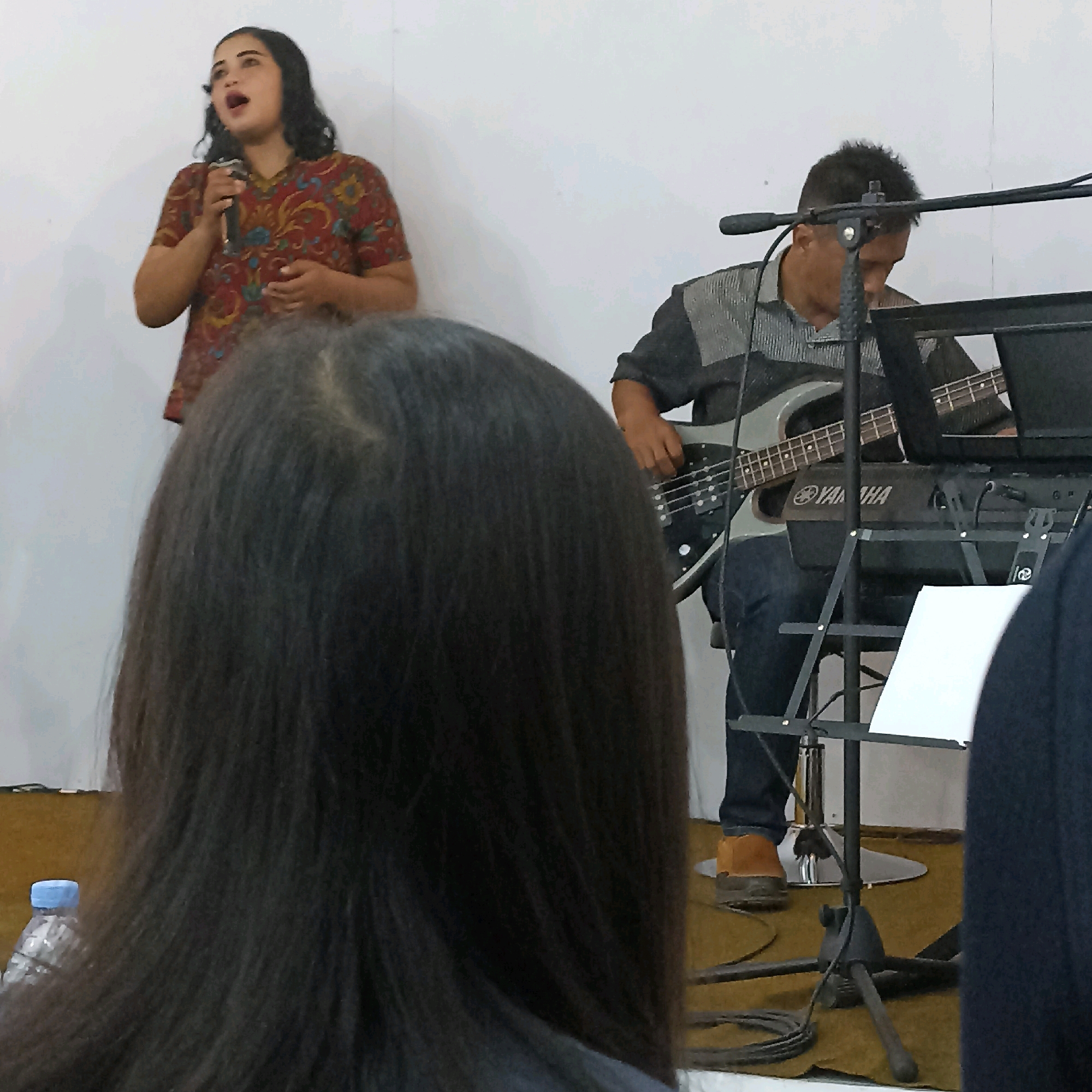 The image depicts a live musical performance by a band in a room with a white wall and a light brown floor. In the foreground, the back of a person's head with long, dark hair is visible, partially obscuring the view. To the left, a woman with dark, curly hair is singing into a microphone, wearing a colorful, patterned shirt. To the right, a man with short dark hair is playing a silver bass guitar and a keyboard with the brand name "YAMAHA" visible. He is wearing a dark, striped shirt and dark pants, with brown shoes. A microphone stand with a music sheet is positioned in front of him. A partially visible bottle of water with a blue cap is on the left side of the foreground. The scene suggests a performance or rehearsal space, possibly a small venue or studio in Magelang, Indonesia, given the context.