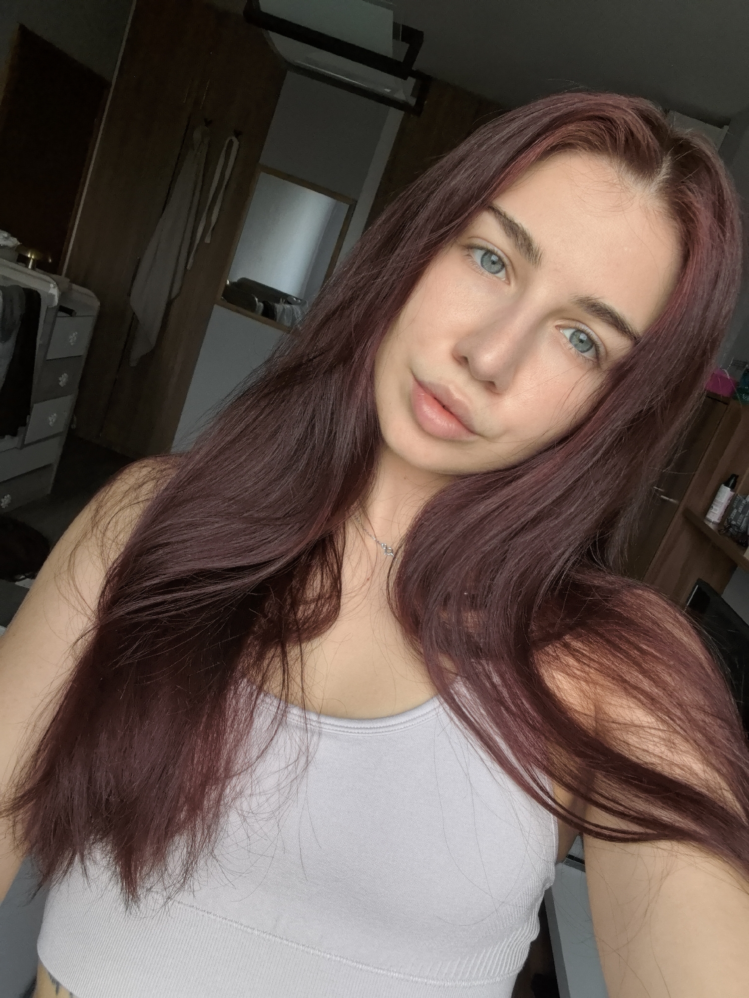 This is a close-up selfie of a young woman with fair skin and striking blue-green eyes, looking directly into the camera. Her long, dark red (auburn or burgundy) hair is parted slightly and flows over her shoulders and chest. She has a neutral to slightly serene expression on her face, with full lips. She is wearing a light grey or pale lavender ribbed tank top and a delicate silver-colored necklace with a small pendant.
The background indicates an indoor setting, likely a bedroom or personal living space. On the left, dark wooden furniture, possibly a dresser or side table with decorative handles, is visible. Further back, another piece of dark wooden furniture, resembling a wardrobe, has a light-colored strap or item hanging on it. To the right, a rectangular mirror with a wooden frame reflects a blurred portion of the room, including what appears to be shelves or additional dark furniture. The lighting is natural but somewhat dim, suggesting an indoor daytime setting without strong direct light. There is no visible text in the image.