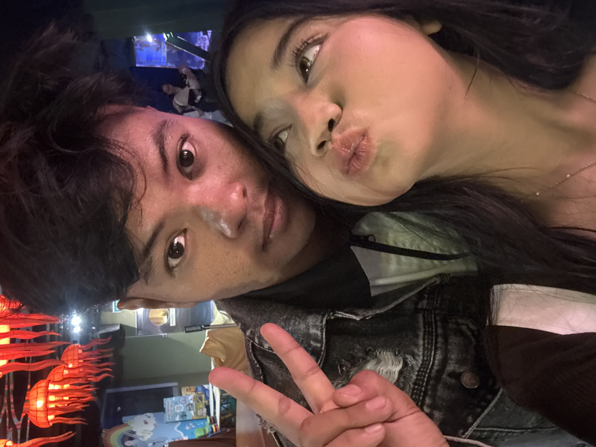 A selfie features two individuals in an indoor, dimly lit, marine-themed environment, consistent with a venue like the Jakarta Aquarium & Safari in West Jakarta, Indonesia. In the immediate foreground, a female with dark hair and a medium skin tone looks upwards, pouting her lips into a 'kiss' pose. She wears a dark top and a delicate gold-colored necklace. Behind her, a male with dark hair and a medium-dark skin tone displays a peace sign with his left hand, looking directly at the camera. The background is characterized by numerous red, illuminated, jellyfish-shaped lanterns suspended from the ceiling. To the left, a portion of a wall mural depicting a rainbow and clouds is visible. To the right, a blue-lit display or aquarium is present, with a blurred human figure partially visible behind it. A light brown bag rests on a surface in the mid-background.