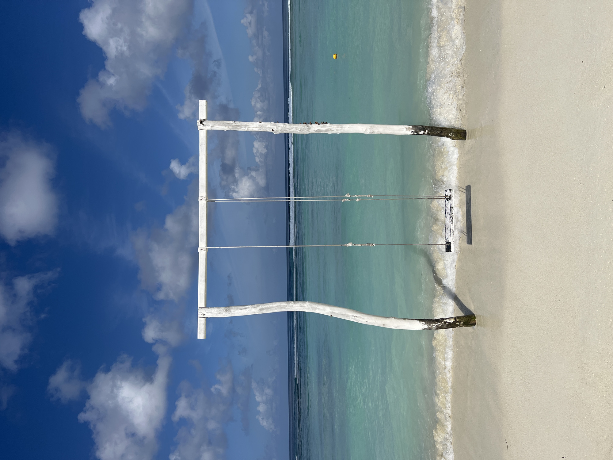 A wooden swing set with a white horizontal bar and two white, gnarled posts is positioned on a sandy beach. The swing seat is made of weathered white wood. The turquoise ocean stretches out behind the swing set, with gentle waves lapping at the shore. In the distance, the horizon is visible, where the ocean meets a bright blue sky filled with white, fluffy clouds. A yellow buoy floats on the water to the right of the swing set. The scene appears to be in the city of Malé, Maldives.