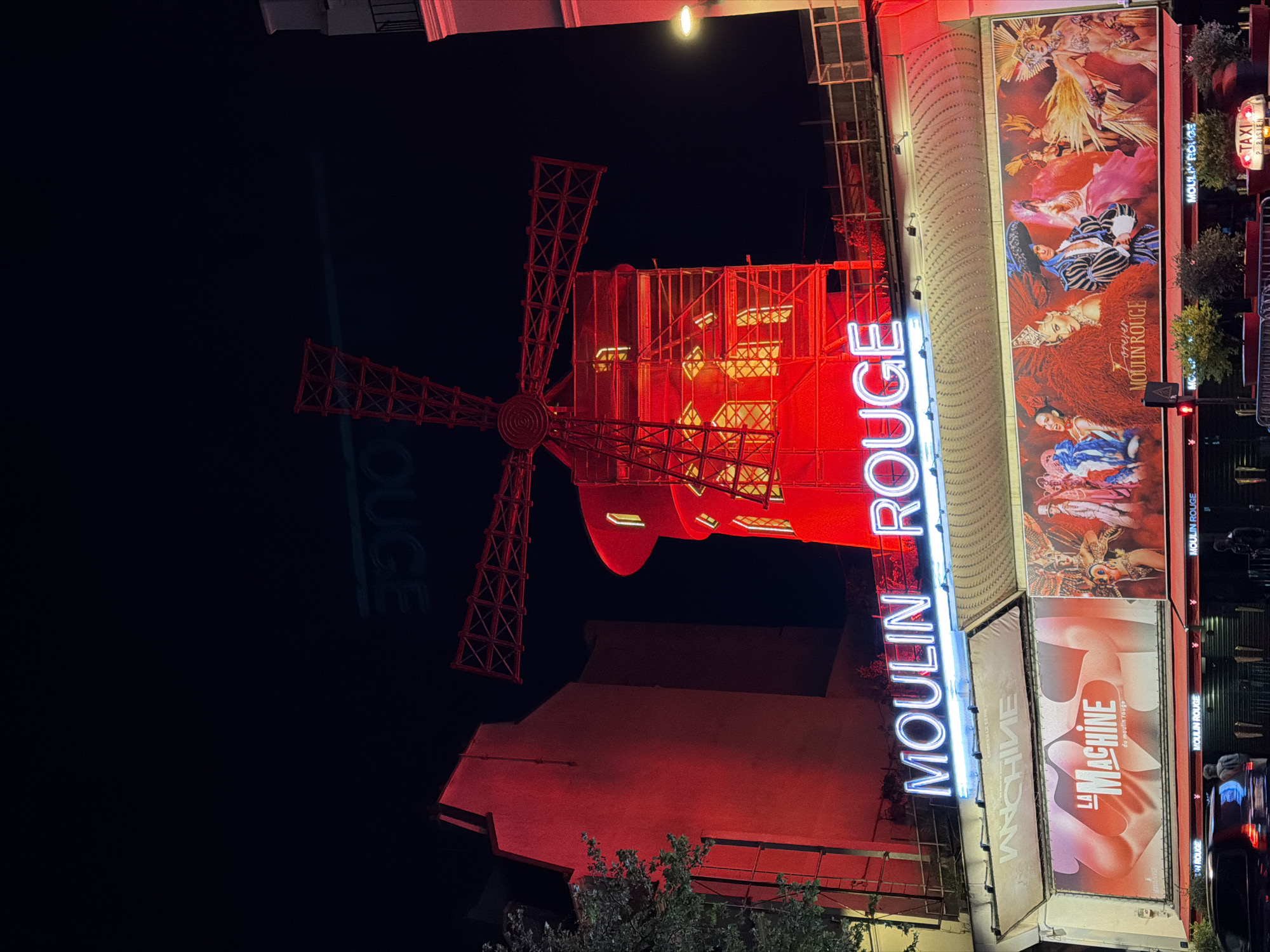 The image depicts the Moulin Rouge in Paris, France, at night. The iconic red windmill is prominently featured, illuminated with red lights. The name "MOULIN ROUGE" is spelled out in bright neon letters. Adjacent to the windmill, a series of posters and billboards are displayed, advertising the Moulin Rouge shows, including "La Machine." The environment is dark, with some street lighting and foliage visible. There is a taxi cab sign visible in the distance.